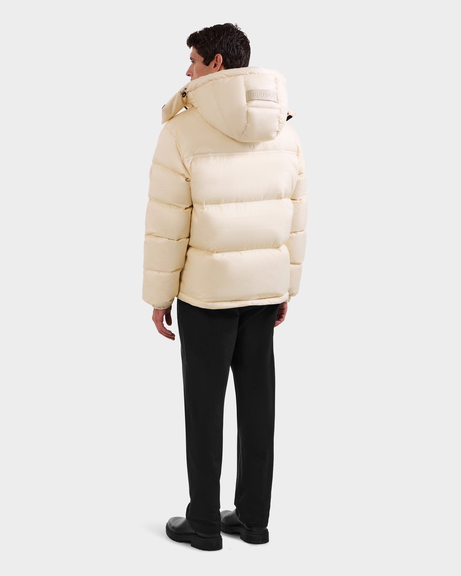 A person faces away, wearing the Rudsak STEFANO - 6125574 Vanilla men’s down puffer jacket with a hood, paired with black pants and shoes, standing against a plain light gray background.