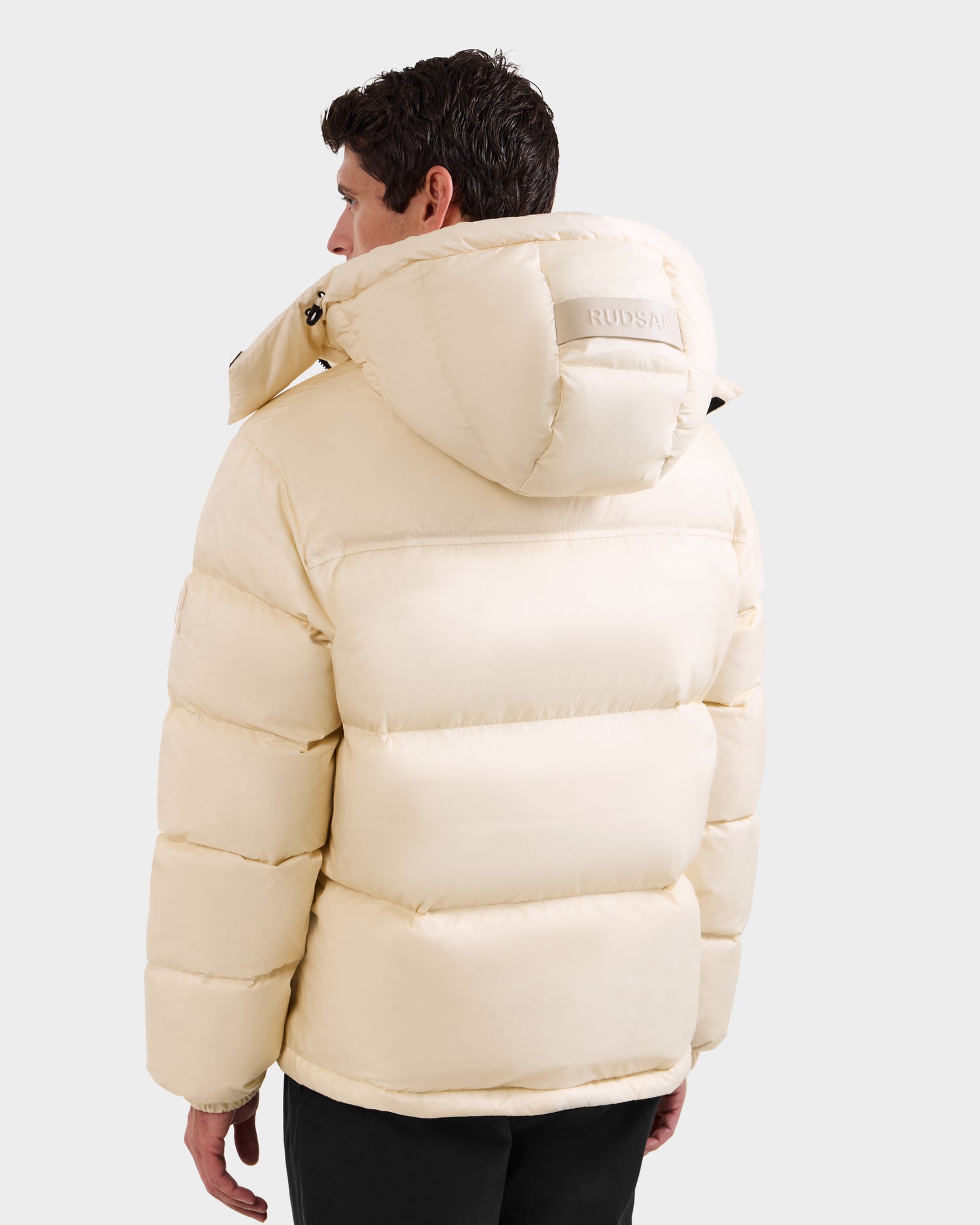 A person is seen from the back wearing Rudsak’s STEFANO - 6125574 Vanilla men’s down puffer jacket with a hood, horizontal quilting, and a patch on the hood, against a plain light background.