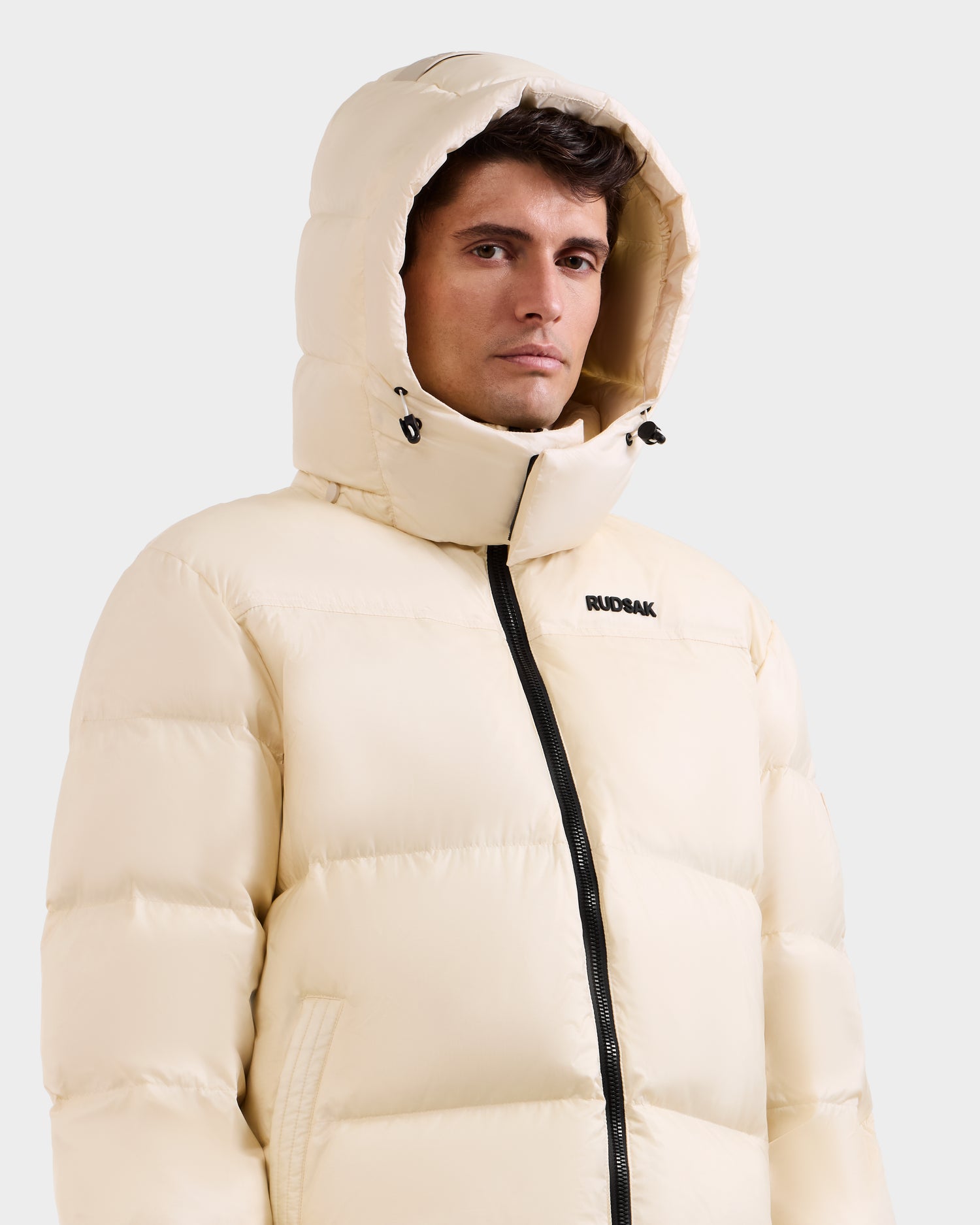 A man wears the Rudsak STEFANO - 6125574 Vanilla men's down puffer jacket with a hood and black zipper, featuring "RUDSAK" on the chest and filled with RDS-certified down. He stands against a plain white background.