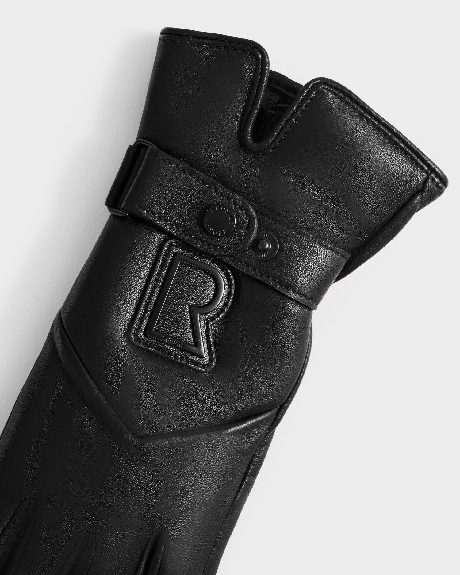 A close-up of the Rudsak STROMBOLI - 8424510 Black women’s leather glove, showcasing a raised R logo, snap button strap, and stitched detailing on a light gray background.