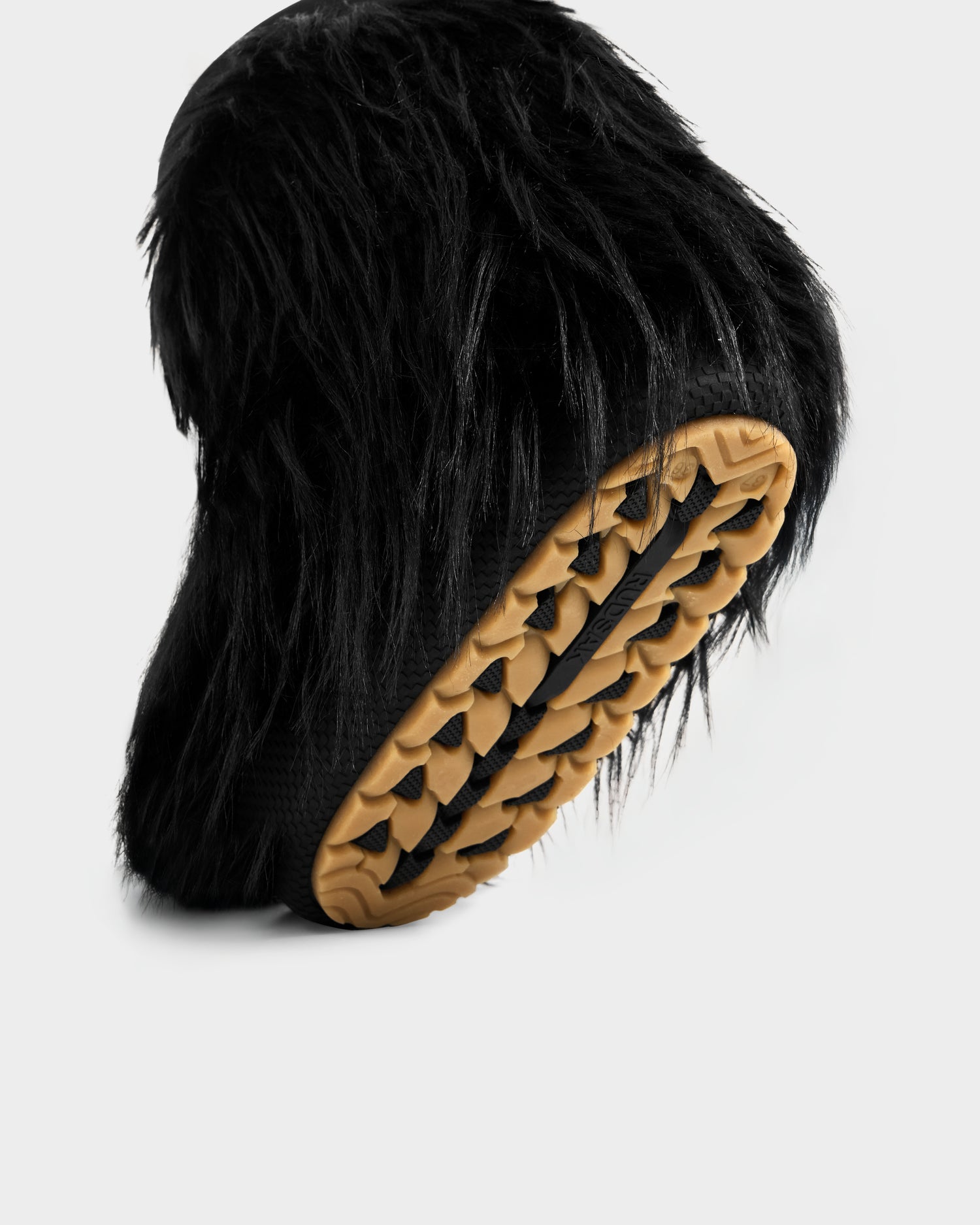 The SUZIE G - 8225545 Black by Rudsak is a women’s moon boot featuring long black faux fur and a textured tan rubber sole, shown against a white background—ideal as bold winter statement boots.