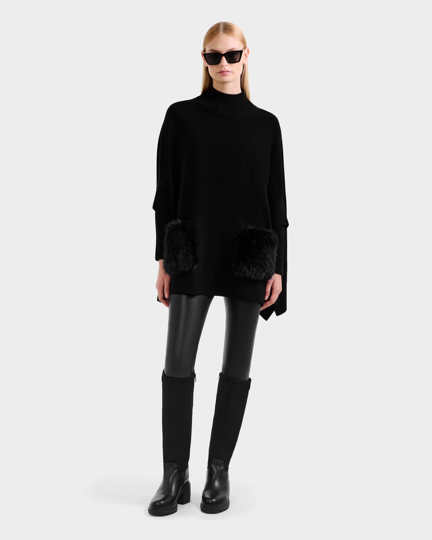 A woman in black sunglasses models the Rudsak SWOON - 8525672 Black layered knit sweater poncho with faux fur pockets, styled with black leather pants and knee-high boots, against a light gray background.