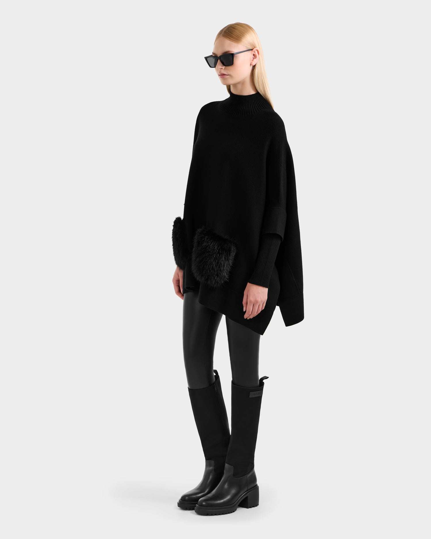 A woman models the Rudsak SWOON - 8525672 Black sweater poncho with faux fur pockets, styled with black sunglasses, black leggings, and knee-high boots against a plain light gray background.