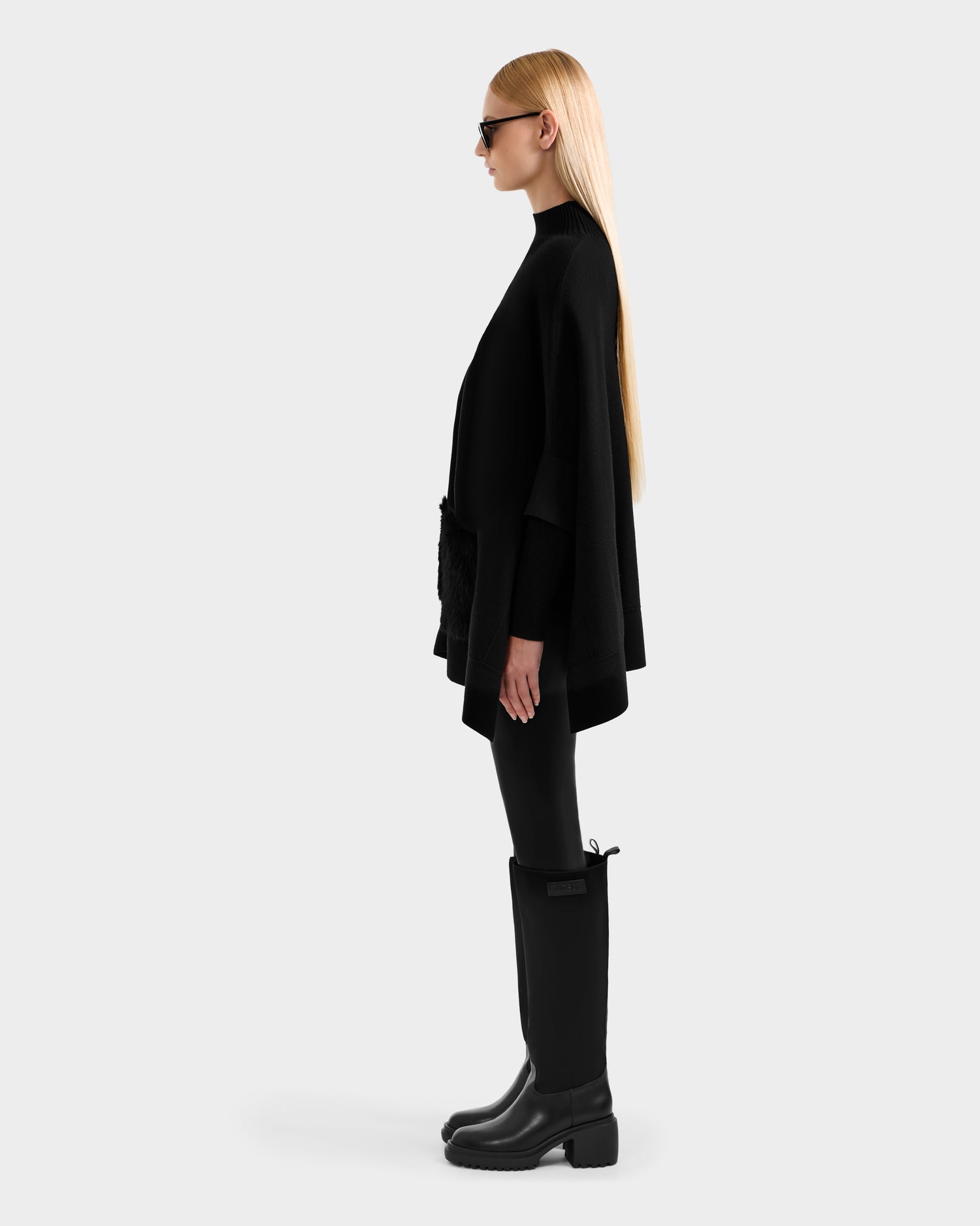 A woman with long, straight blonde hair stands in profile against a plain background, wearing sunglasses and the Rudsak SWOON - 8525672 Black sweater poncho with faux fur pocket details, black leggings, and black knee-high boots.