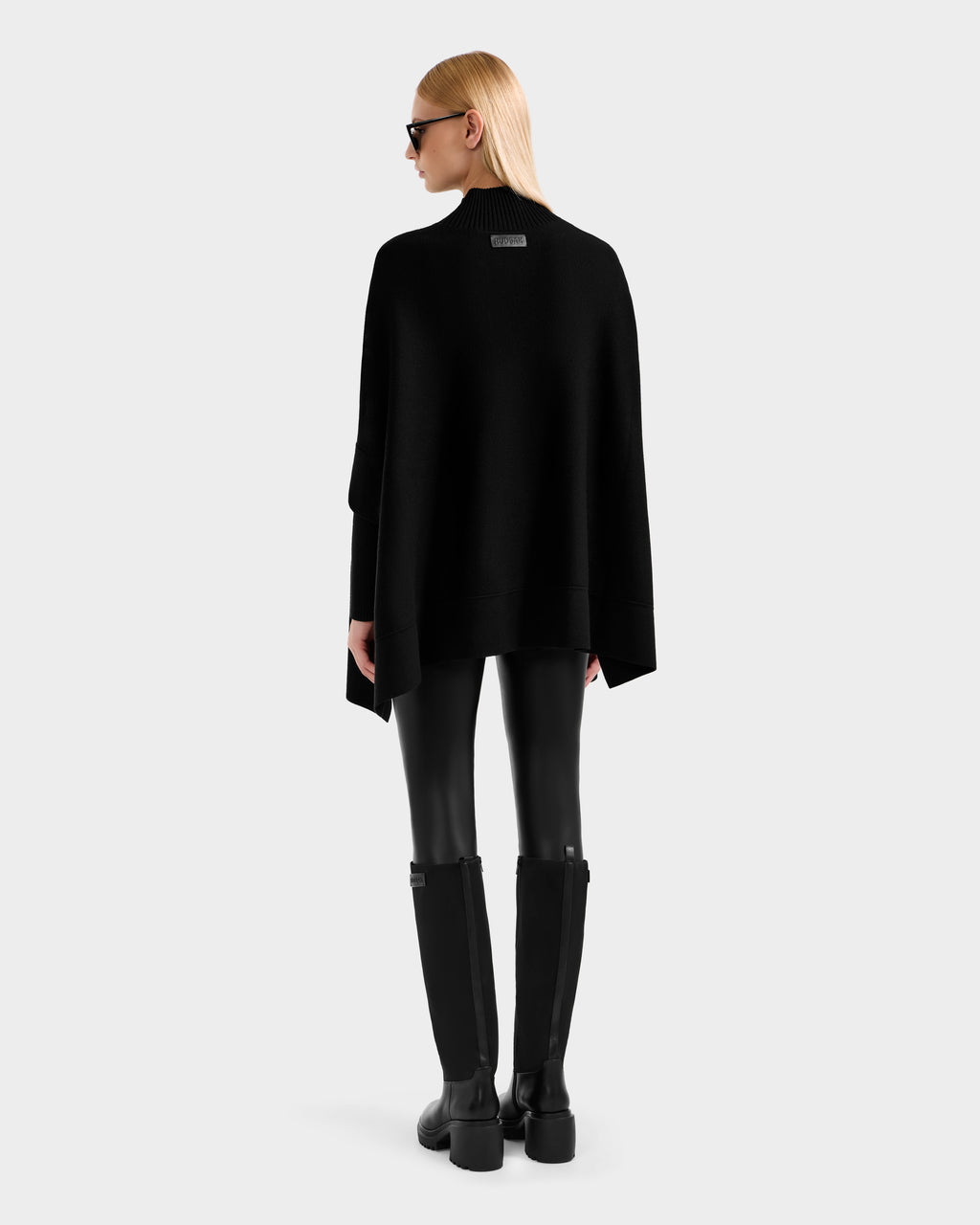 A person with straight blonde hair in sunglasses models the Rudsak SWOON - 8525672 Black knit poncho with faux fur pockets, paired with black leather pants and tall boots, standing against a light gray background.