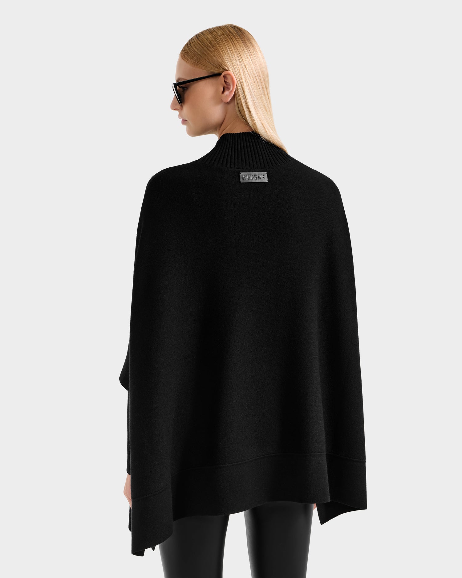 A person with straight blonde hair, black sunglasses, and black pants is seen from behind wearing the Rudsak SWOON - 8525672 Black layered knit turtleneck cape, with a visible BUDDHA label, against a light gray background.