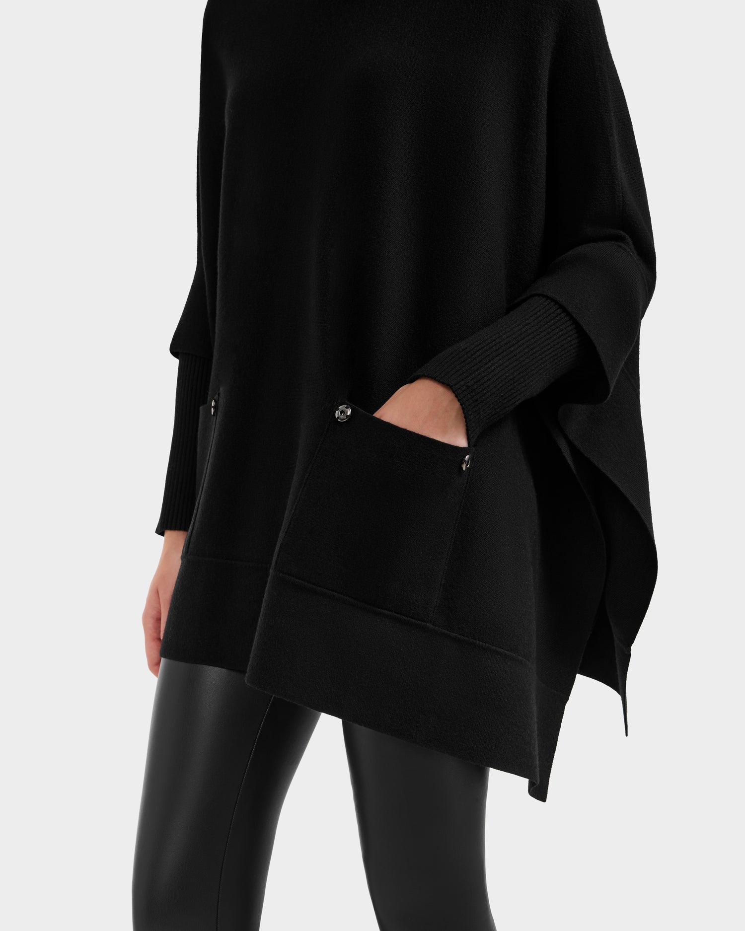 Someone wears the Rudsak SWOON - 8525672 Black sweater poncho, featuring front pockets and a loose, layered knit drape. The lower face and body are visible against a plain light background, styled with black faux leather leggings.
