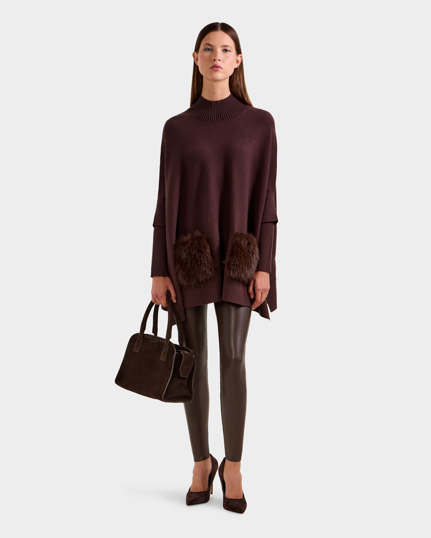 A woman wears the Rudsak SWOON - 8525672 Mocha sweater poncho with faux fur pockets, paired with brown leather leggings, brown heels, and a brown handbag against a plain light background.