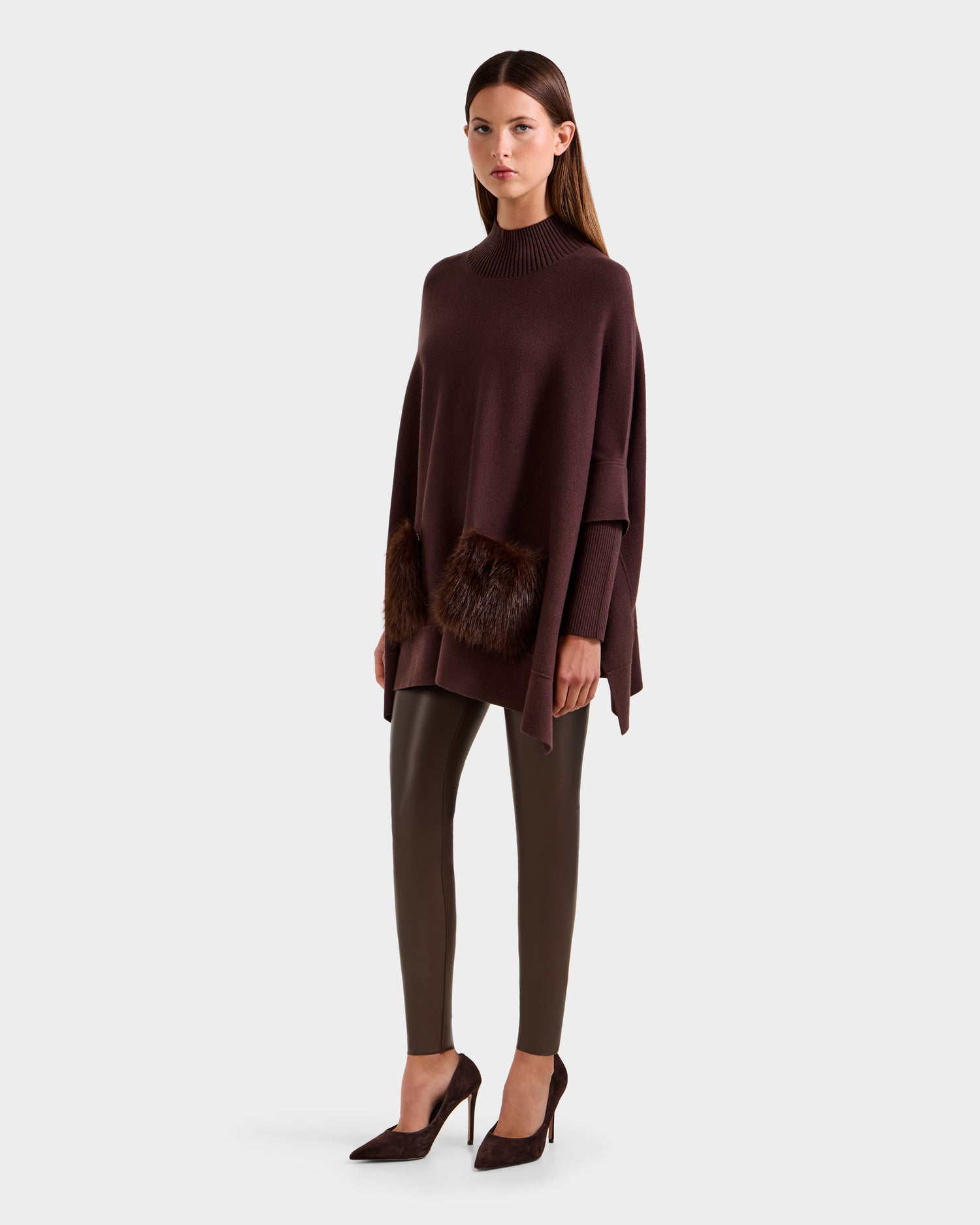 A woman faces forward against a white background, wearing Rudsak's SWOON - 8525672 Mocha sweater poncho with faux fur pockets, paired with brown leggings and dark brown heels.