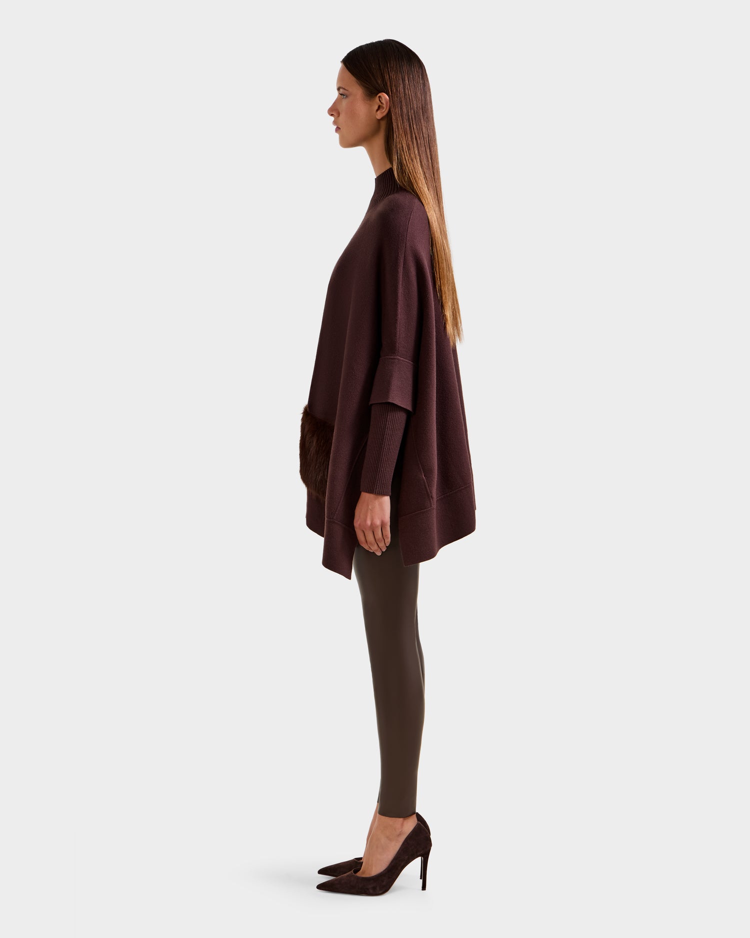 A woman in profile wears the Rudsak SWOON - 8525672 Mocha loose dark brown sweater poncho with faux fur pocket accents, styled with fitted brown leggings and dark heels. Her straight hair is worn down, highlighting modern layering.