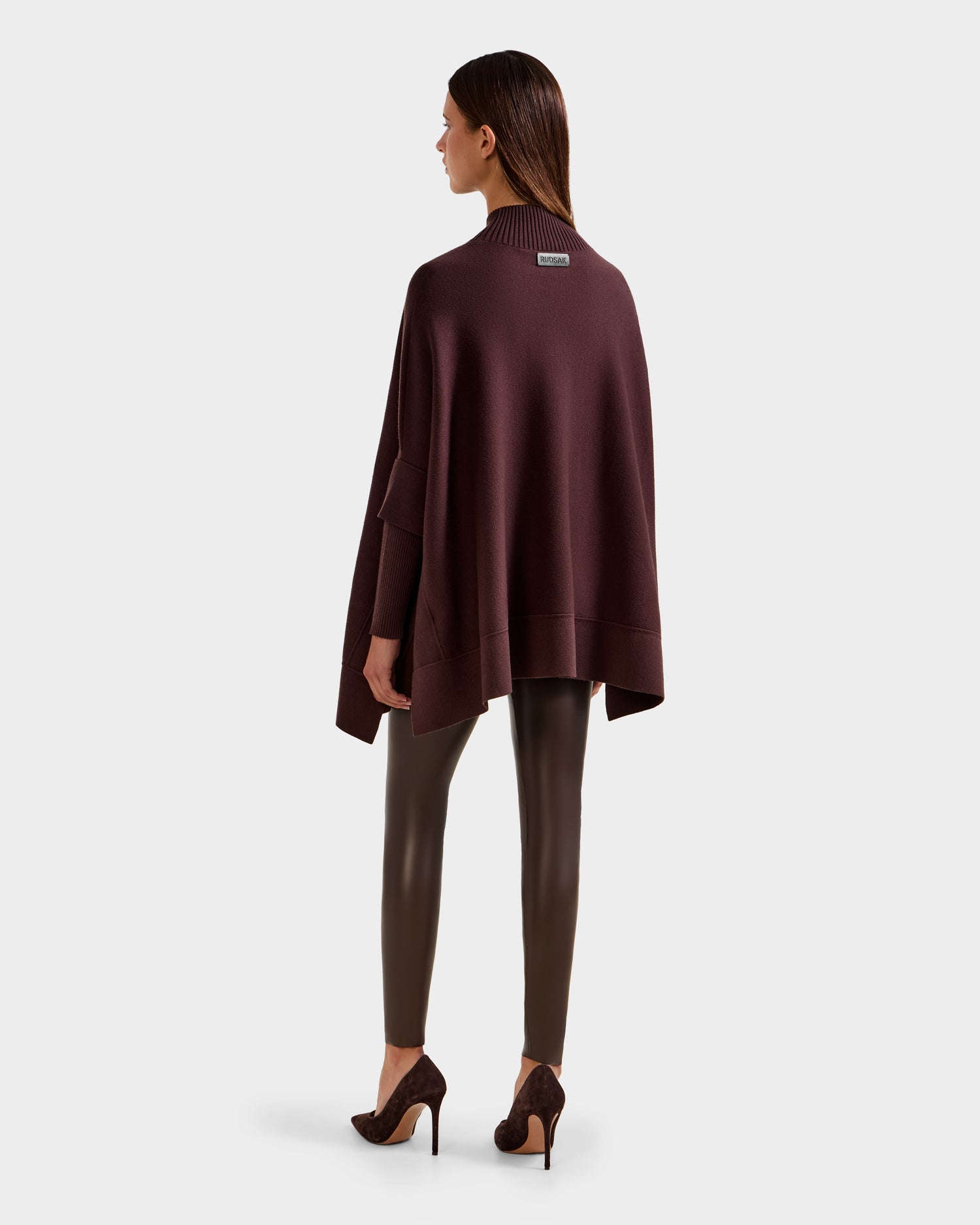 A woman faces away, wearing Rudsak's SWOON - 8525672 Mocha loose dark maroon sweater poncho, styled with dark brown leggings and brown high-heeled shoes against a plain light gray background.