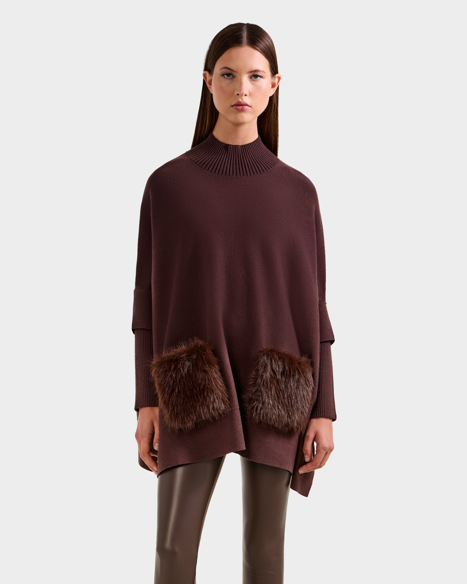 A woman models the Rudsak SWOON - 8525672 Mocha poncho, featuring a ribbed turtleneck and faux fur pockets, paired with brown leather pants against a plain light background for a modern layered look.
