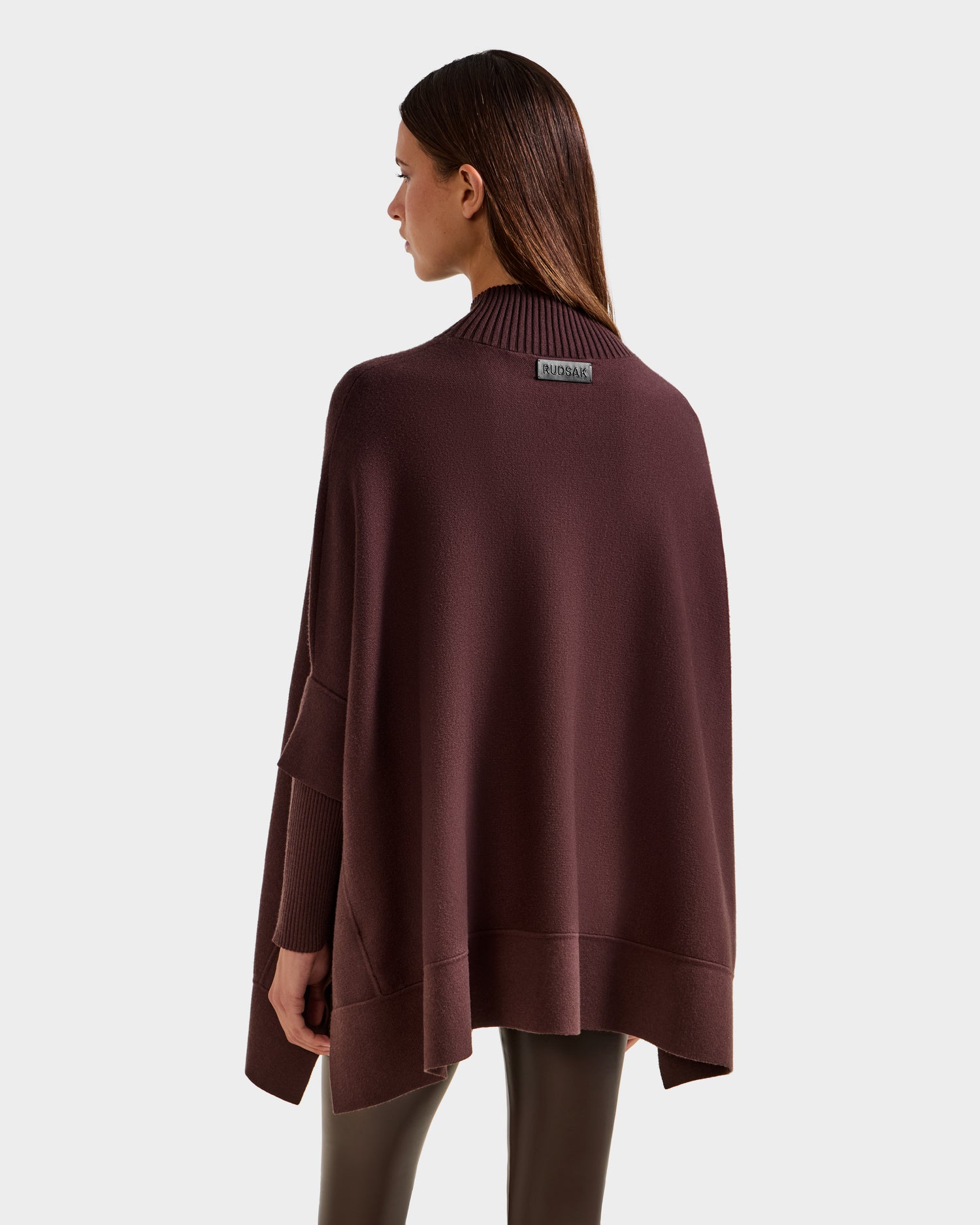 A woman with straight brown hair, seen from the back, wears the Rudsak SWOON - 8525672 Mocha sweater poncho with ribbed cuffs and collar—a modern layering piece—featuring a small label on the back and paired with dark leggings.