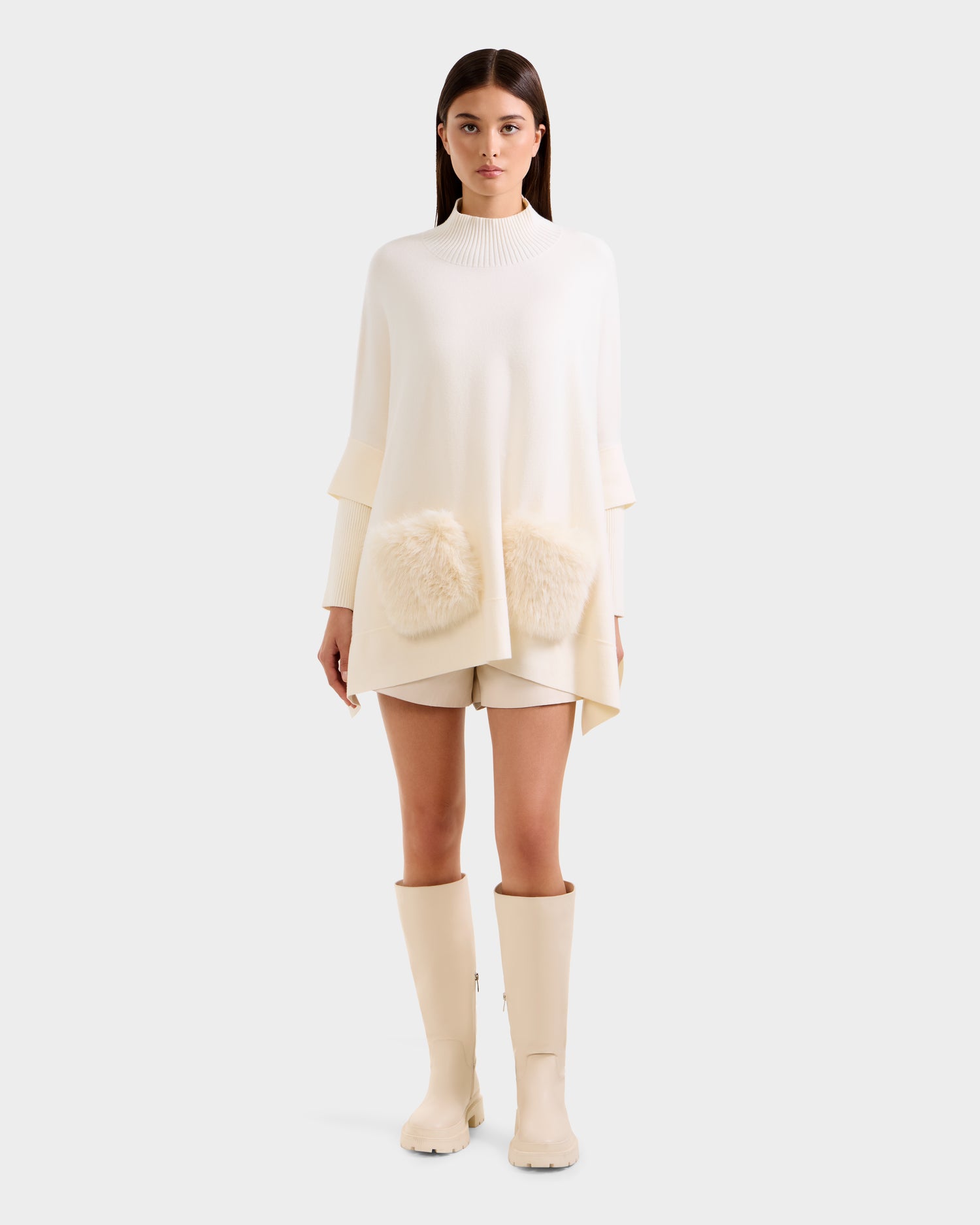 A woman faces forward in Rudsak’s SWOON - 8525672 Vanilla sweater poncho with faux fur pockets, paired with matching shorts and ivory knee-high boots, exemplifying modern layering against a plain light background.