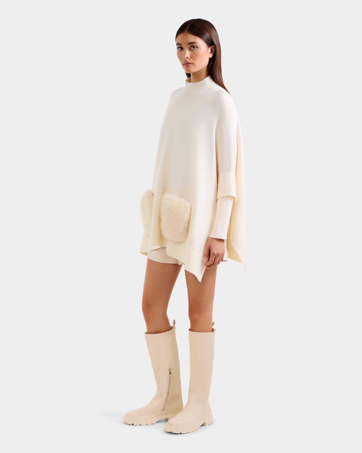 A woman models the Rudsak SWOON - 8525672 Vanilla: an oversized cream sweater poncho with faux fur pockets, paired with matching cream shorts and knee-high boots, styled against a plain light background.