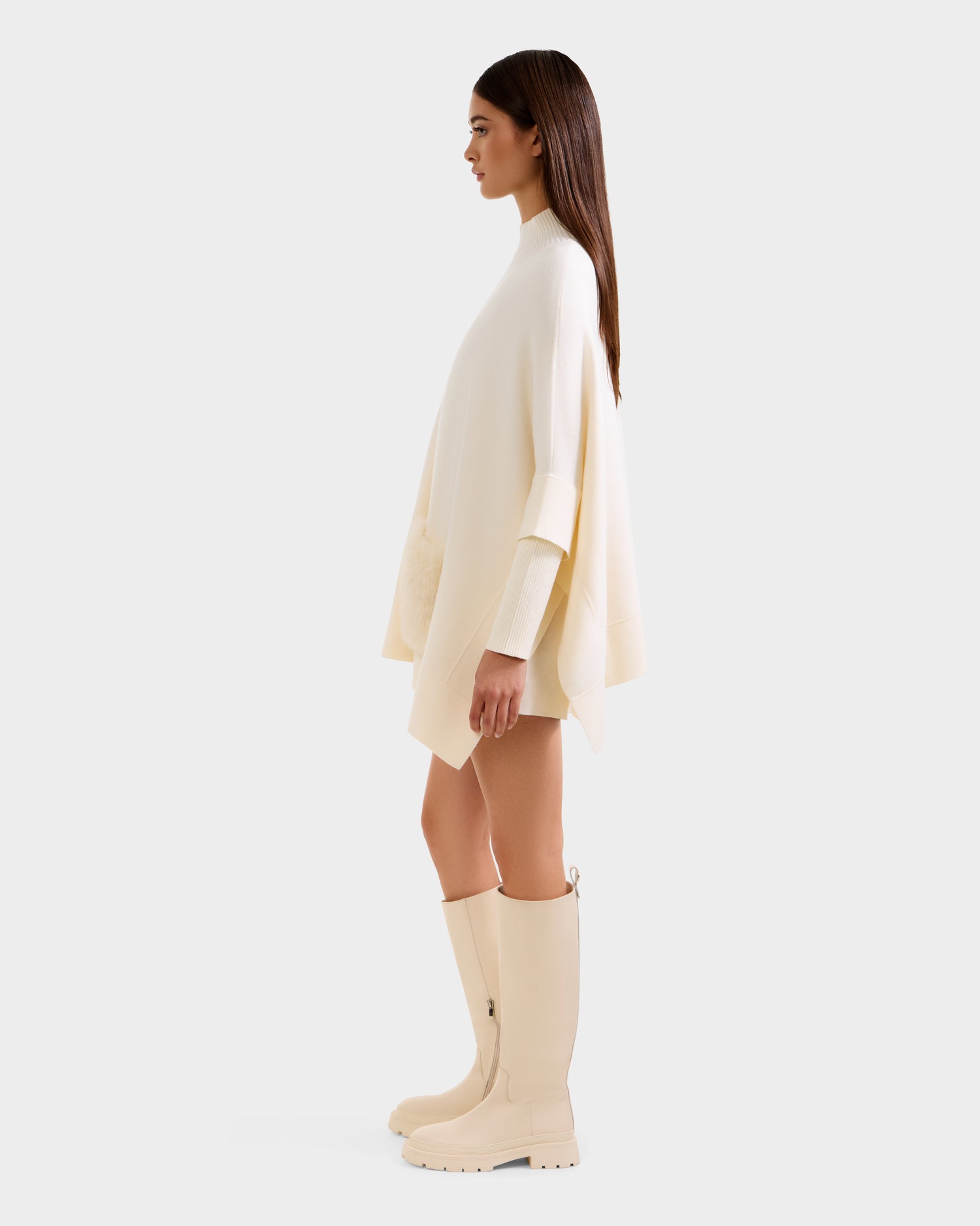 A woman in profile wears the Rudsak SWOON - 8525672 Vanilla sweater poncho, matching mini skirt, and knee-high boots. Her long brown hair is loose, highlighting the modern, minimalist monochrome look against a plain background.