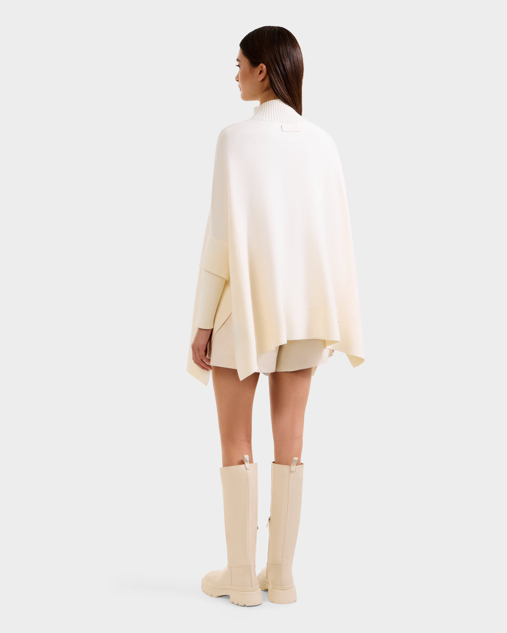 A woman faces away in a modern look, wearing Rudsak's SWOON - 8525672 Vanilla ivory sweater poncho with faux fur pocket, paired with a cream mini skirt and off-white knee-high boots, set against a plain light background.