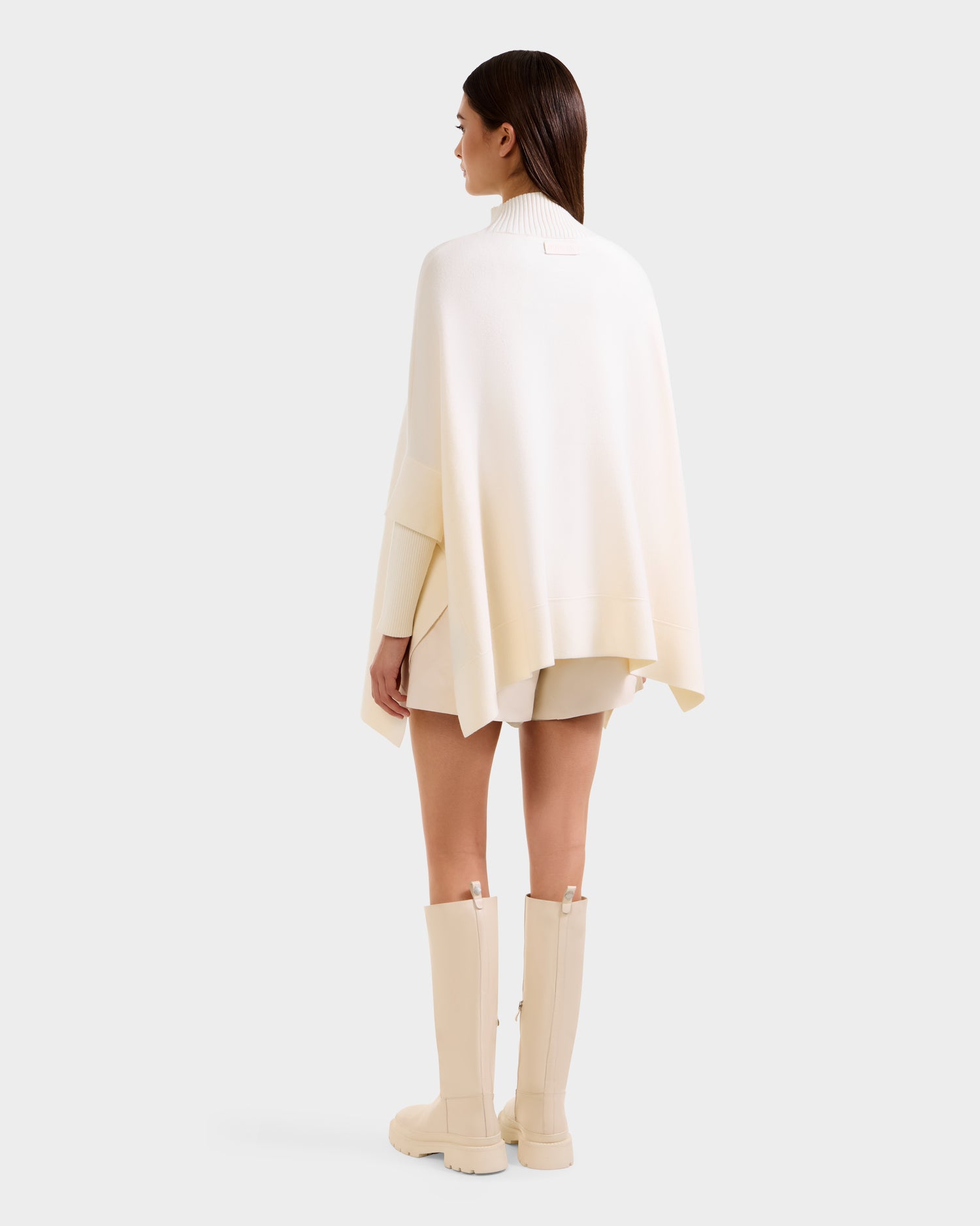 A woman faces away in a modern look, wearing Rudsak's SWOON - 8525672 Vanilla ivory sweater poncho with faux fur pocket, paired with a cream mini skirt and off-white knee-high boots, set against a plain light background.