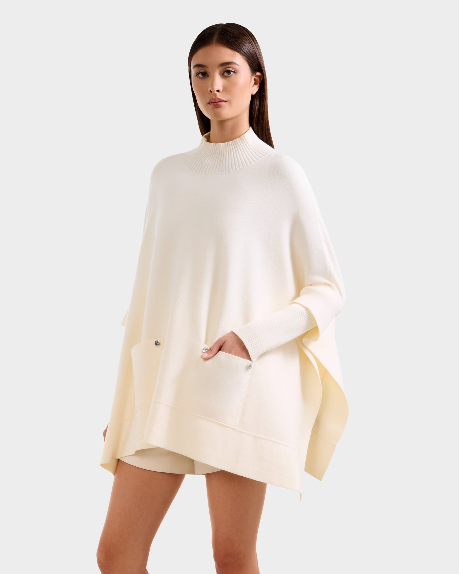 A woman stands facing forward against a light gray background, showcasing the SWOON - 8525672 Vanilla sweater poncho by Rudsak—a loose, cream-colored piece with a ribbed mock neck and front pockets for effortless modern layering.