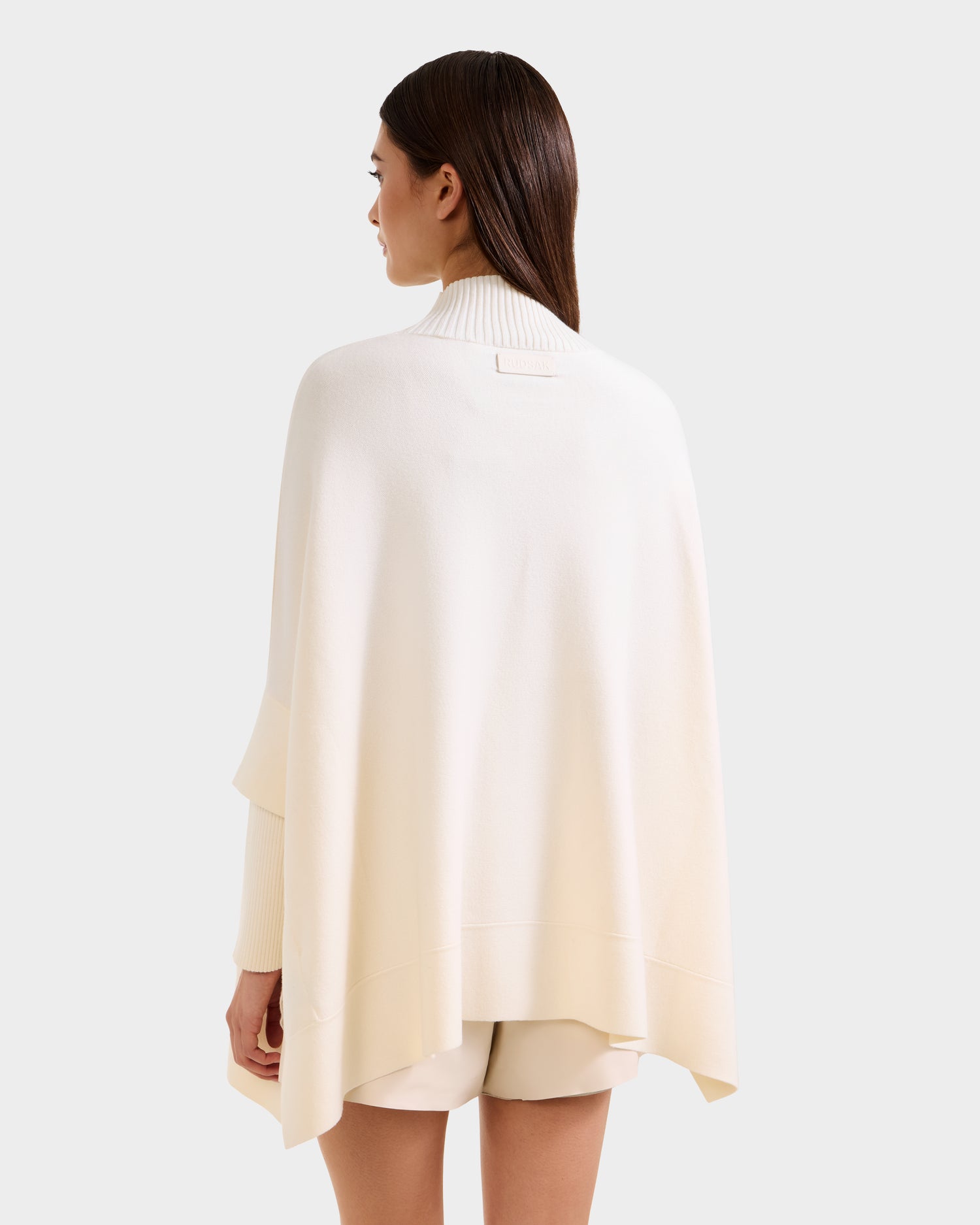 A woman with straight brown hair wears the Rudsak SWOON - 8525672 Vanilla off-white sweater poncho and matching shorts, standing facing away against a plain light background.