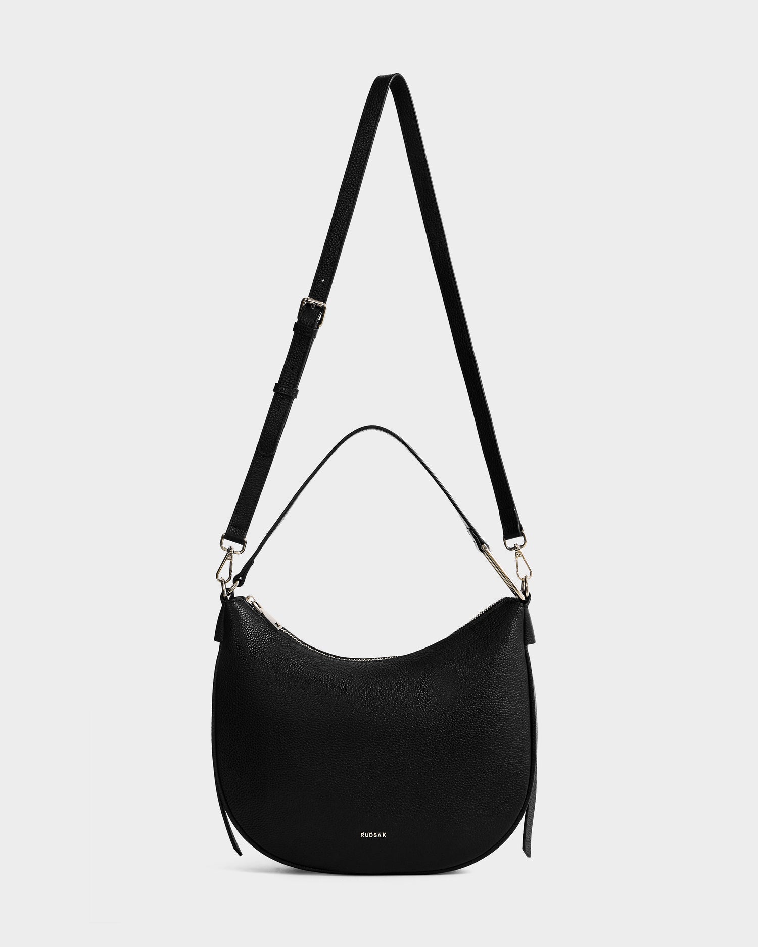 The SYLVIA - L325515 Black by Rudsak is a crescent-shaped women's leather shoulder bag with gold-tone hardware, an adjustable long strap, and short handle, shown against a plain white background.