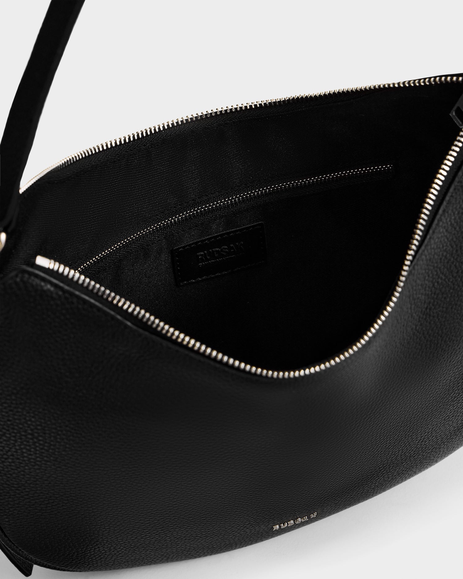 Close-up of an open black women’s leather shoulder bag, SYLVIA - L325515 Black by Rudsak, showing a visible zipper and inner pocket. The brand name is embossed on a leather tag inside, with a plain light-colored background.