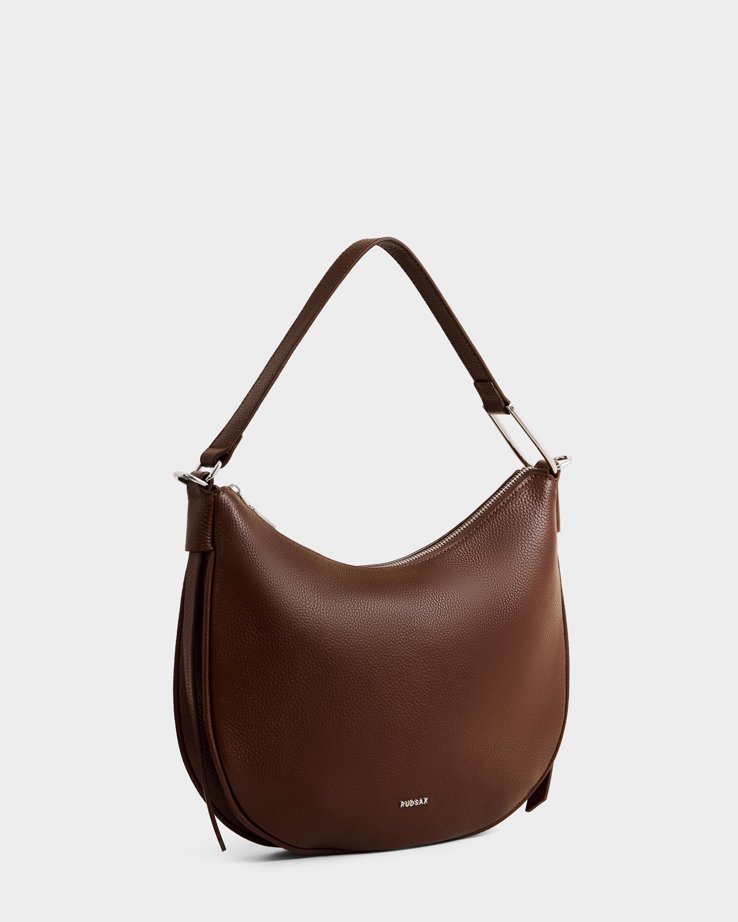 The Rudsak SYLVIA - L325515 Mocha is a women's brown leather shoulder bag with a single wide strap, silver hardware, subtle front branding, and a crescent silhouette. Photographed against a plain light background.