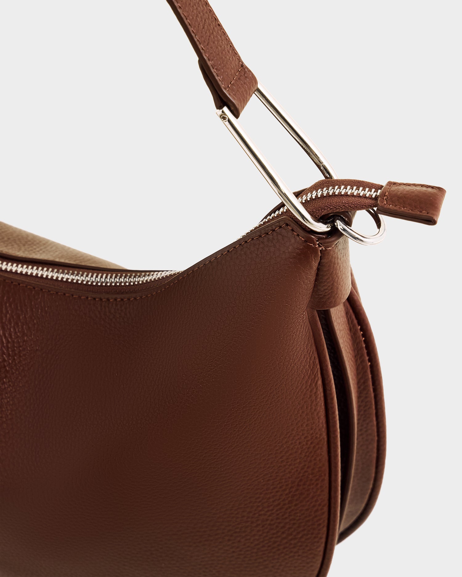 Close-up of the Rudsak SYLVIA - L325515 Mocha leather crossbody bag, featuring a textured brown surface, silver zipper, matching strap with metal ring and rectangular detail, set against a light gray background.