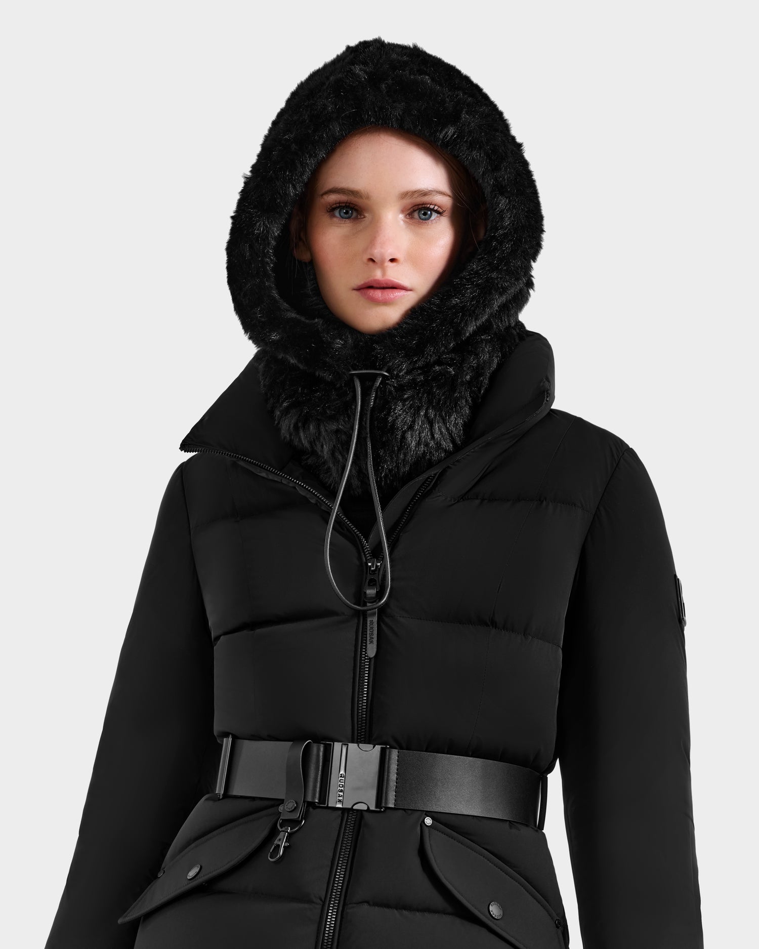 A woman models the Rudsak TARA - 8425025 Black coat, featuring a faux fur-lined hood, zip closure, and belted waist, highlighting timeless winter style against a simple light background.