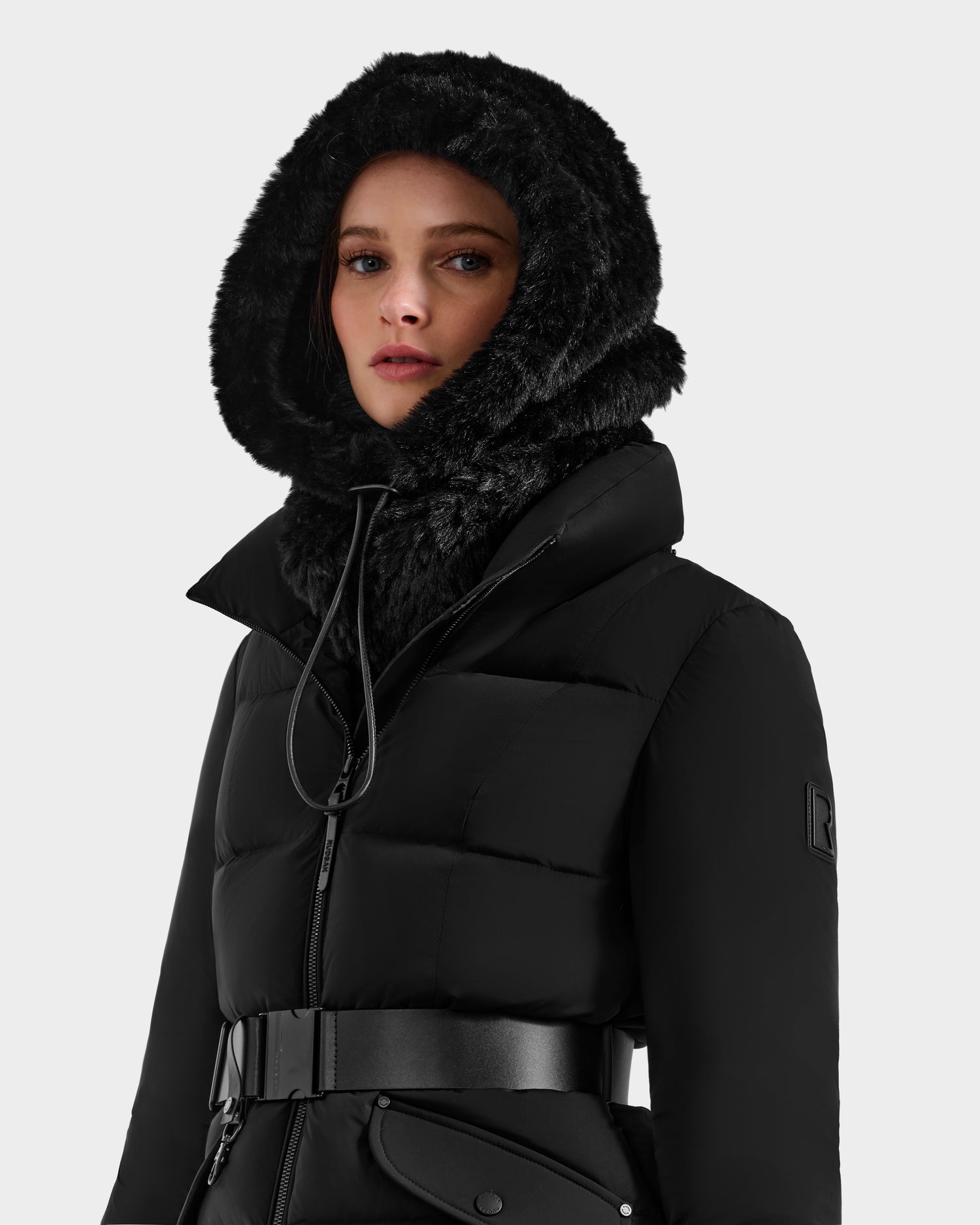 A woman models the Rudsak TARA - 8425025 Black belted puffer coat with a furry hood and a chic RUDSAK balaclava, adding warmth and style as she stands against a plain light background, gazing confidently at the camera.