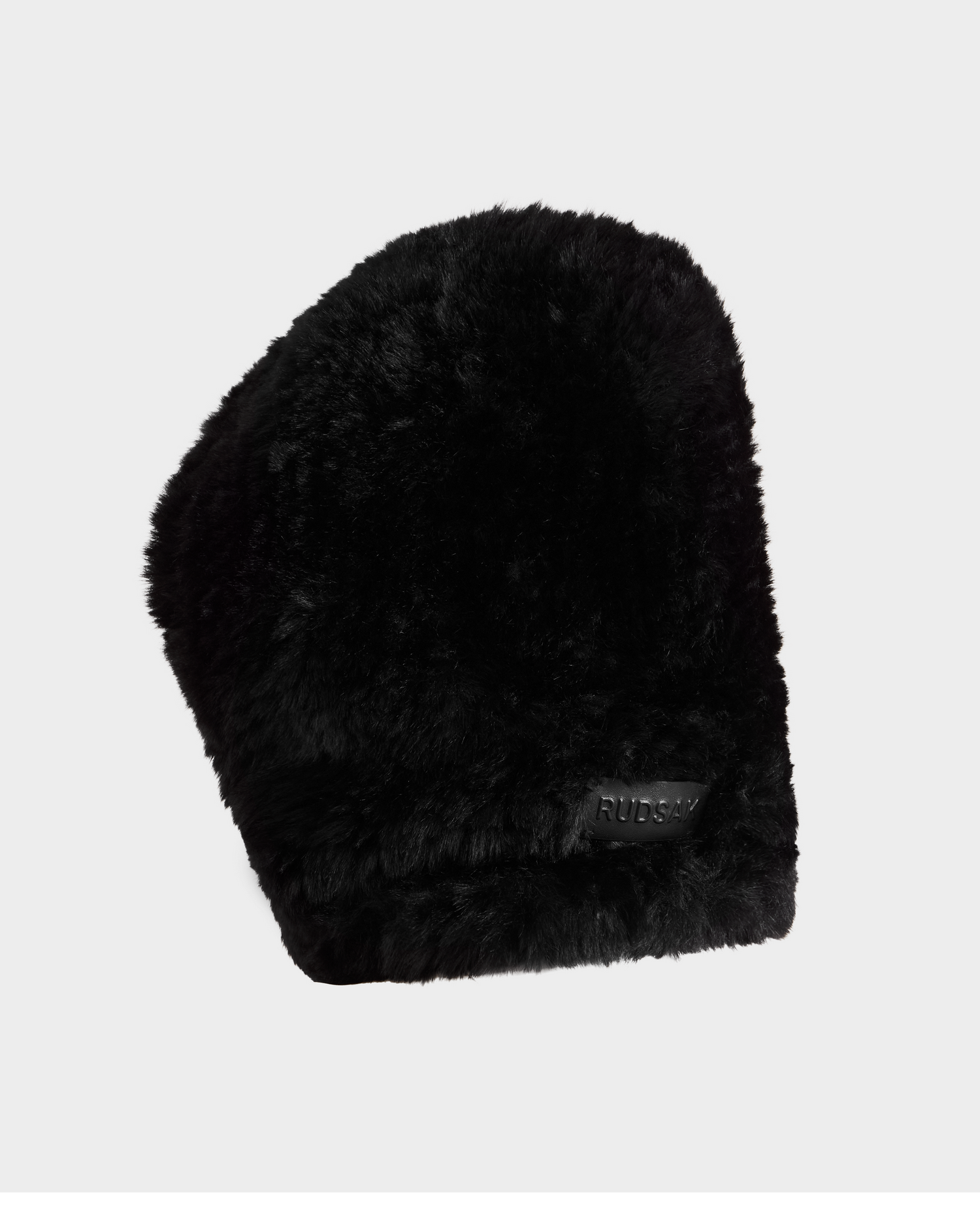 The TARA - 8425025 Black by Rudsak is a black faux fur winter hat with a slightly angled top and a front patch reading RUBSAY. Perfect for winter style, it’s shown against a plain light gray background.