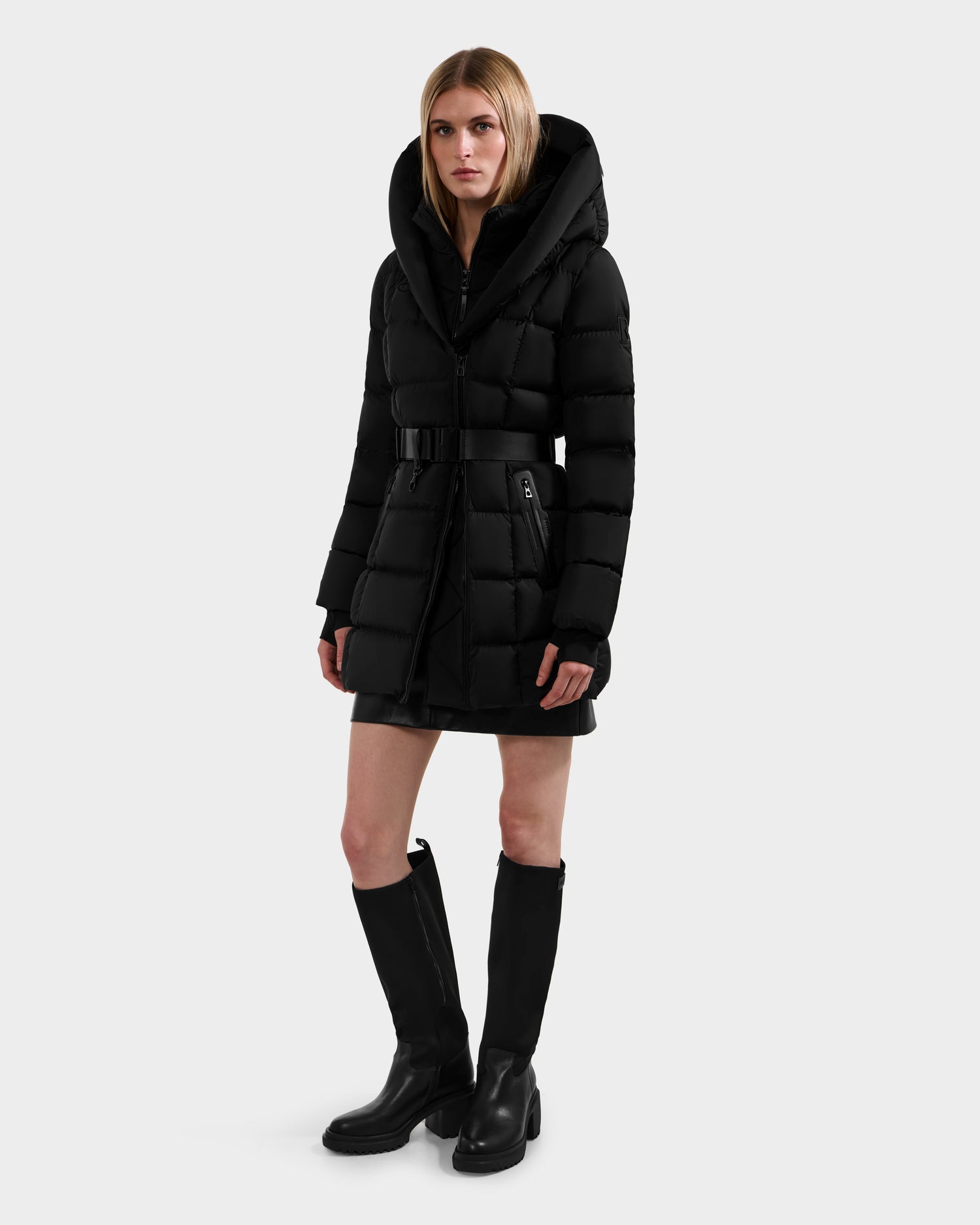 A person poses against a plain light background, wearing the Rudsak TAYLOR M - 8124545 Black quilted puffer jacket with a large hood and black knee-high boots, maintaining a neutral expression.