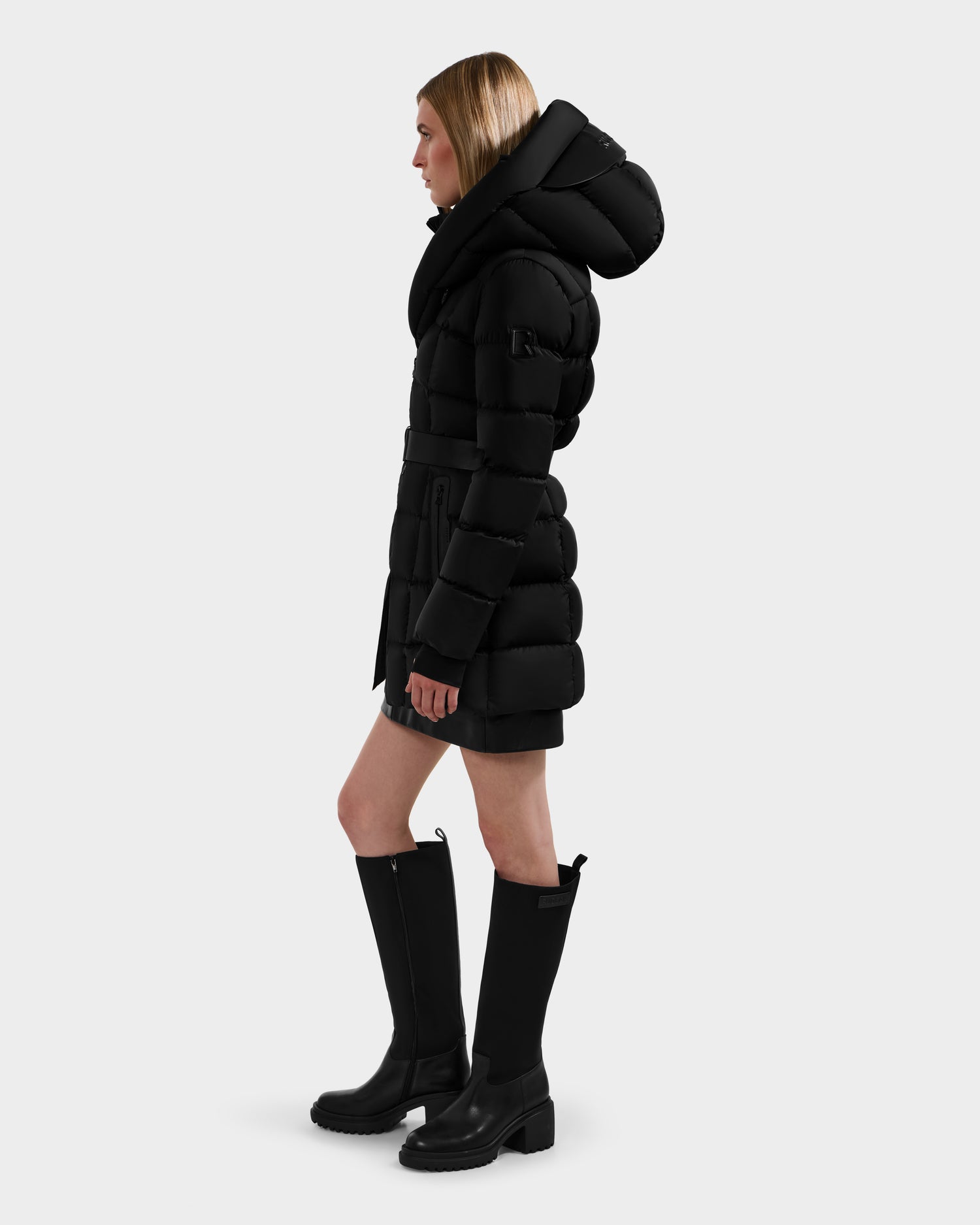 A woman in profile wears the Rudsak TAYLOR M - 8124545 Black down puffer jacket with a large hood and black knee-high boots, standing against a plain light gray background.