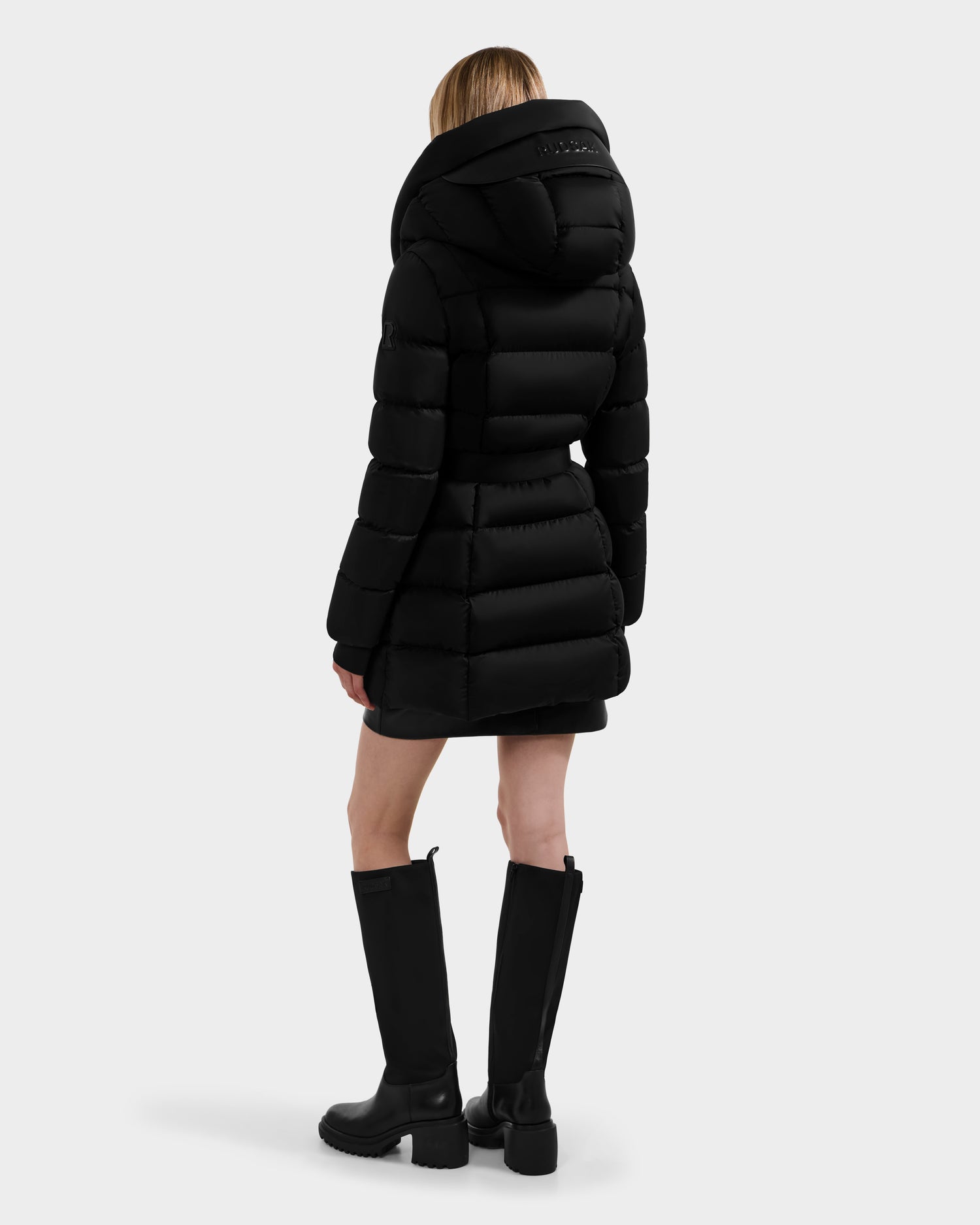 A person models the Rudsak TAYLOR M - 8124545 Black jacket, featuring a large hood, shown from the back with knee-high black boots against a plain light gray background.