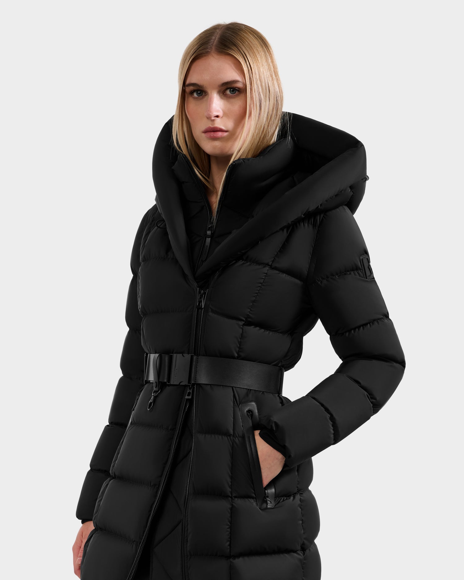 A woman with straight blonde hair models the Rudsak TAYLOR M - 8124545 Black, a long quilted puffer jacket with a wide hood and belted waist, posed slightly left against a plain background with a neutral expression.