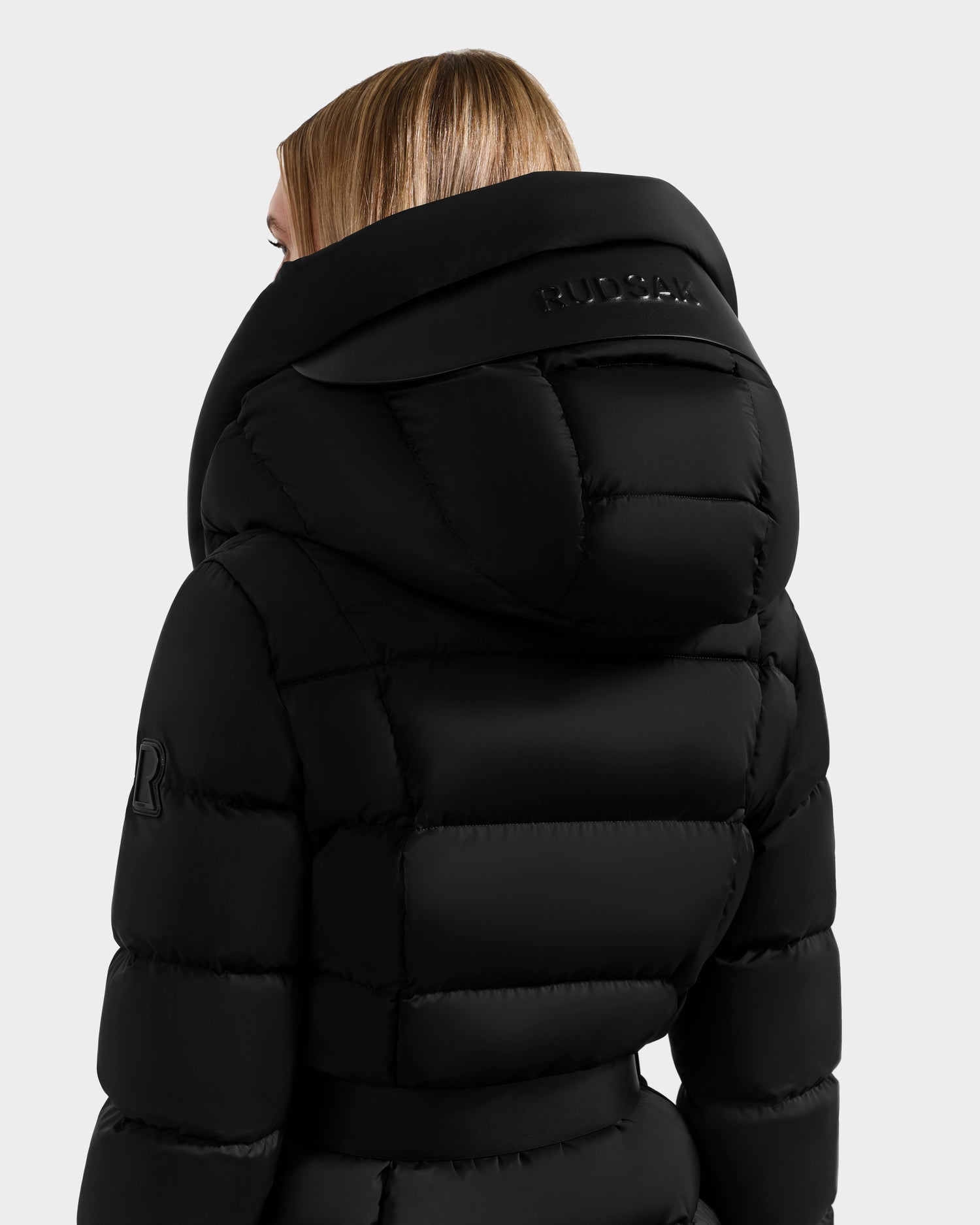 A person with straight blonde hair wears the Rudsak TAYLOR M - 8124545 black quilted puffer jacket, featuring a high collar, large hood with embossed branding, against a plain light gray background.