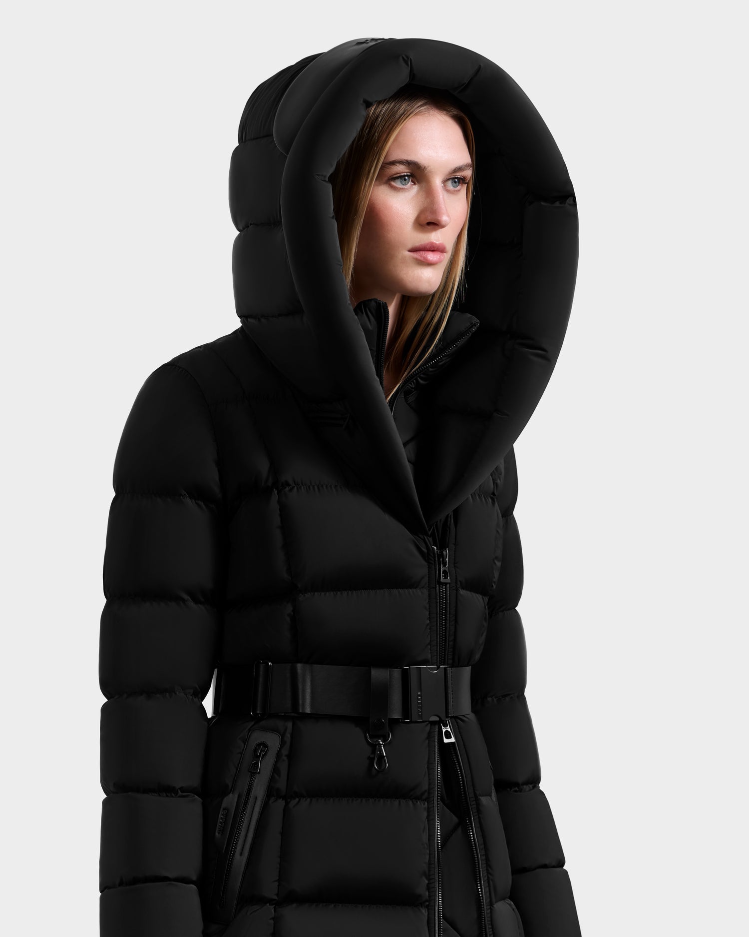 A woman wears the Rudsak TAYLOR M - 8124545 Black quilted winter jacket with a belt and large hood, standing slightly to the side against a plain light background.
