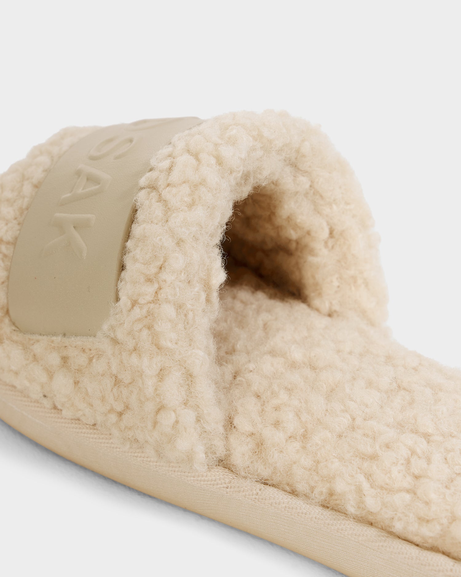 A close-up of the Rudsak TEDDY - L225517 Vanilla women's faux-shearling slipper, featuring a cream-colored textured surface and a wide band with raised letters. Perfect for cozy loungewear, set against a light background.