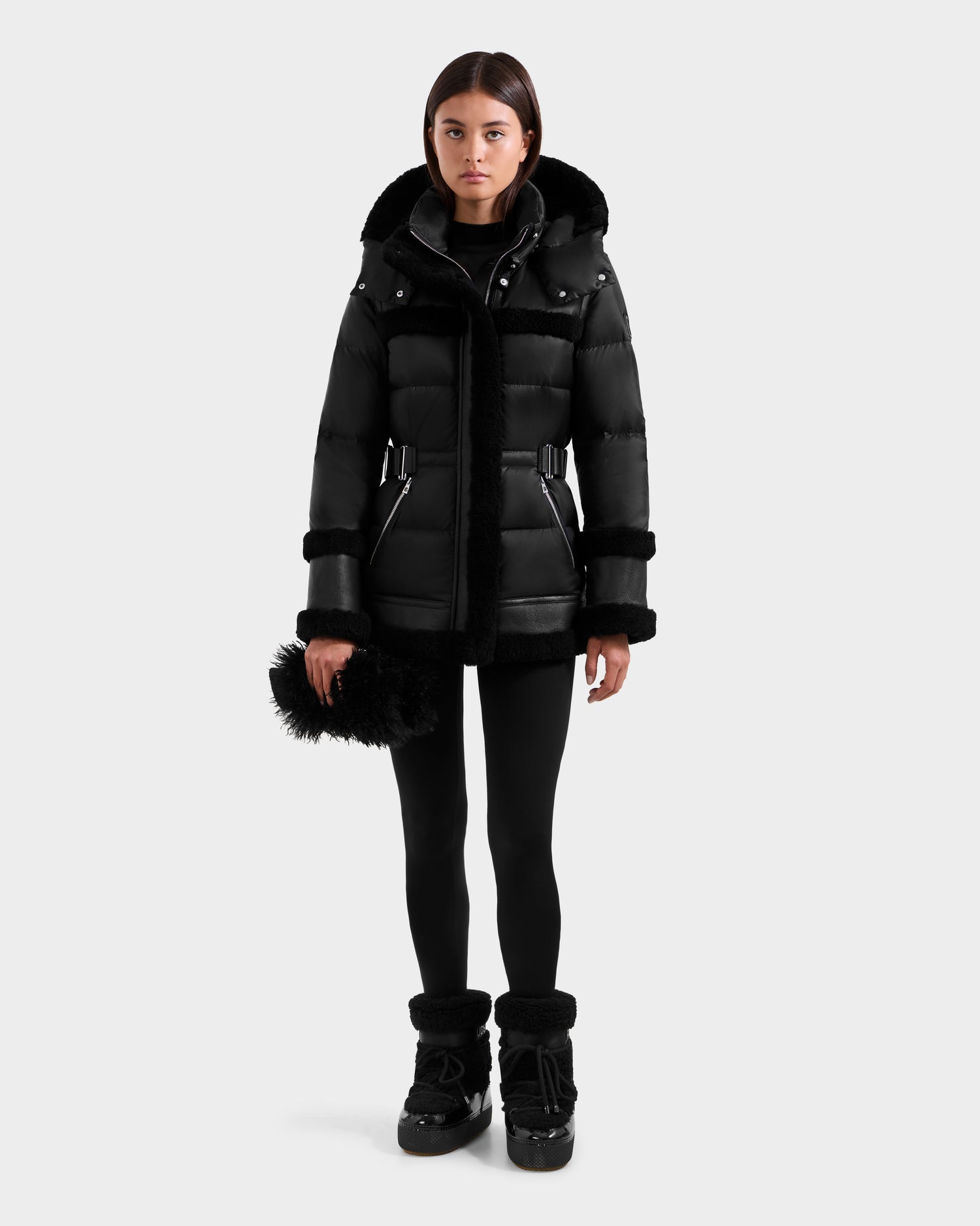 A woman models the Rudsak TESSA - 8125615 Black women’s down puffer with shearling trim, paired with black leggings and fur boots, holding a black fuzzy accessory against a white background—a chic winter look.