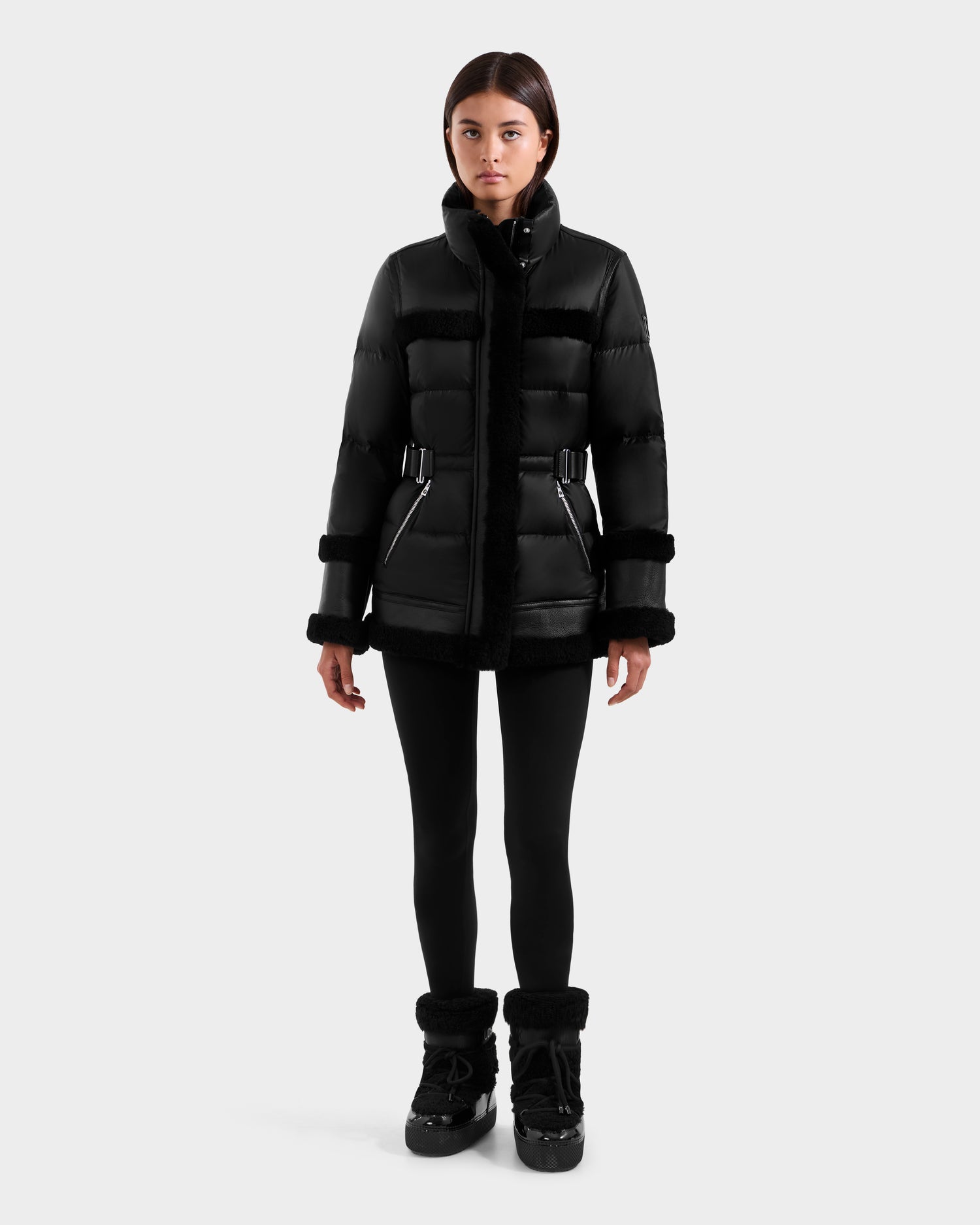 A woman faces forward in the Rudsak TESSA - 8125615 Black down puffer with faux fur trim, paired with black leggings and black winter boots, set against a plain light gray background.