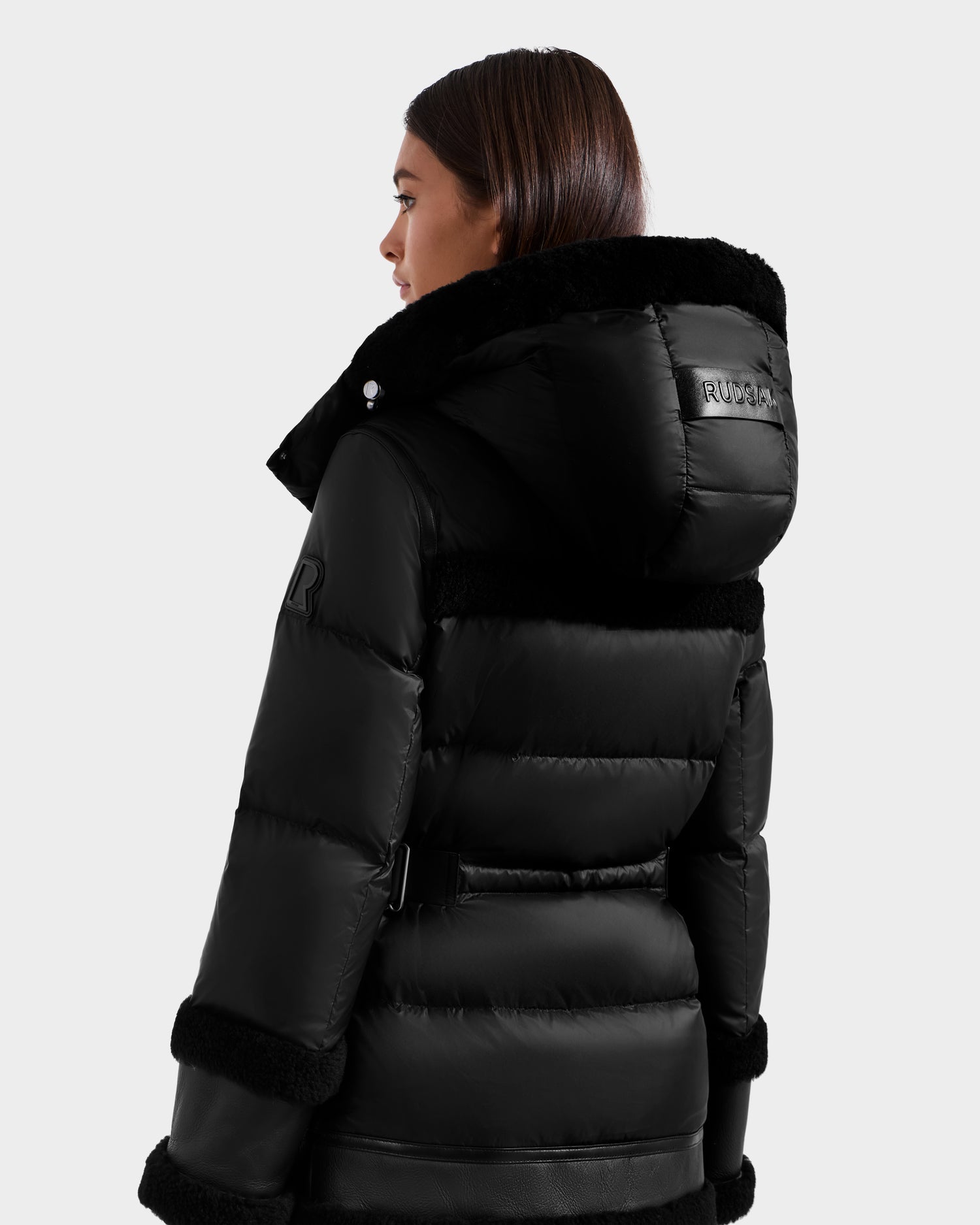 A woman with straight brown hair models the Rudsak TESSA - 8125615 Black women’s down puffer jacket, featuring a hood, shearling trim, and belt detail, while facing away from the camera against a plain light background.
