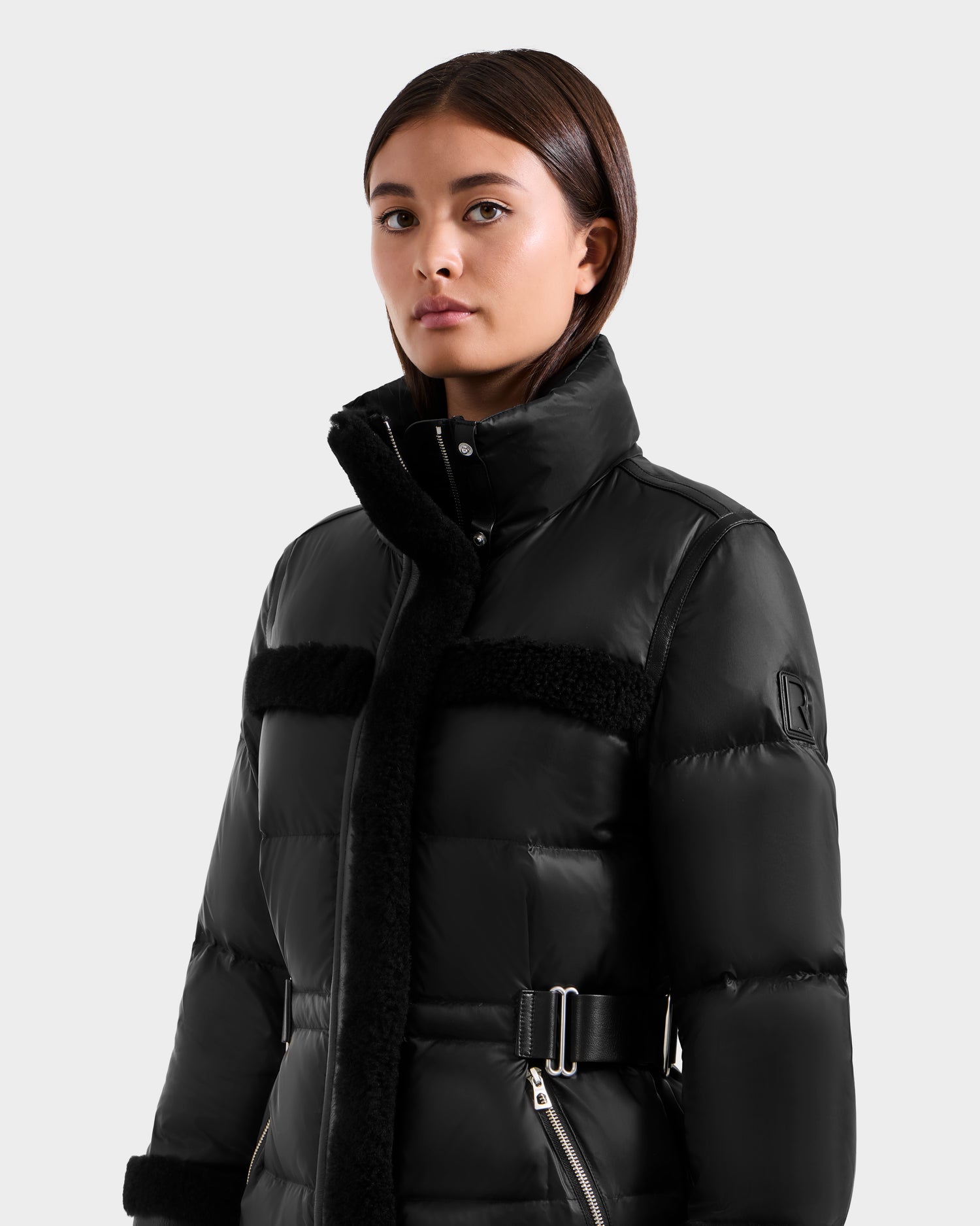 A young woman with straight brown hair wears the Rudsak TESSA - 8125615 Black down puffer coat with a high collar, black shearling trim, and belted waist, standing against a plain light gray background and looking slightly to the side.