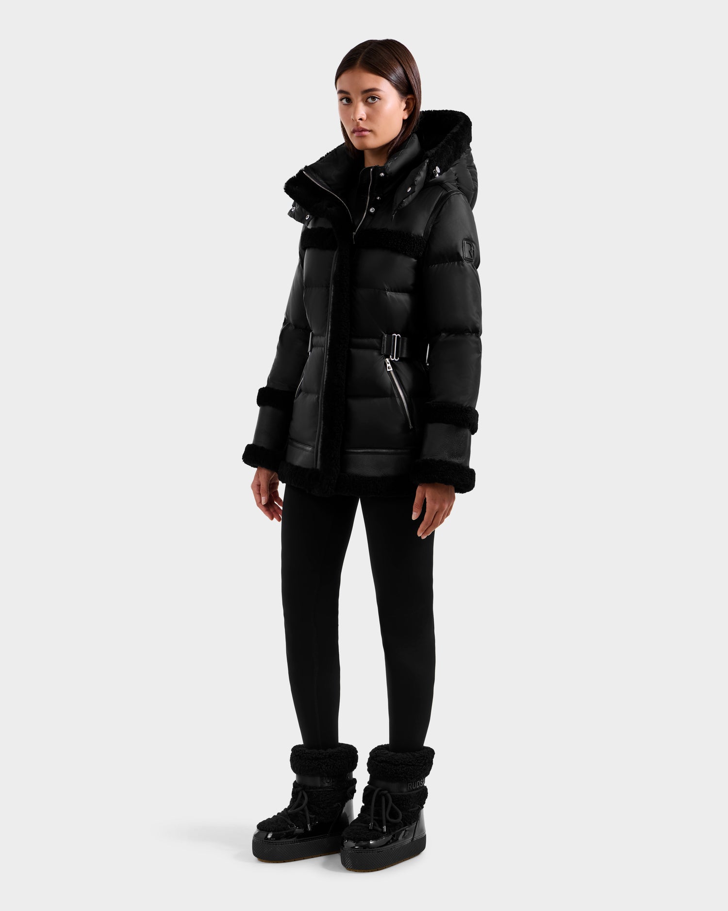 A person stands against a plain background wearing the Rudsak TESSA - 8125615 Black women’s down puffer jacket with a belt and hood, paired with black pants and winter boots. Their hands are relaxed at their sides.