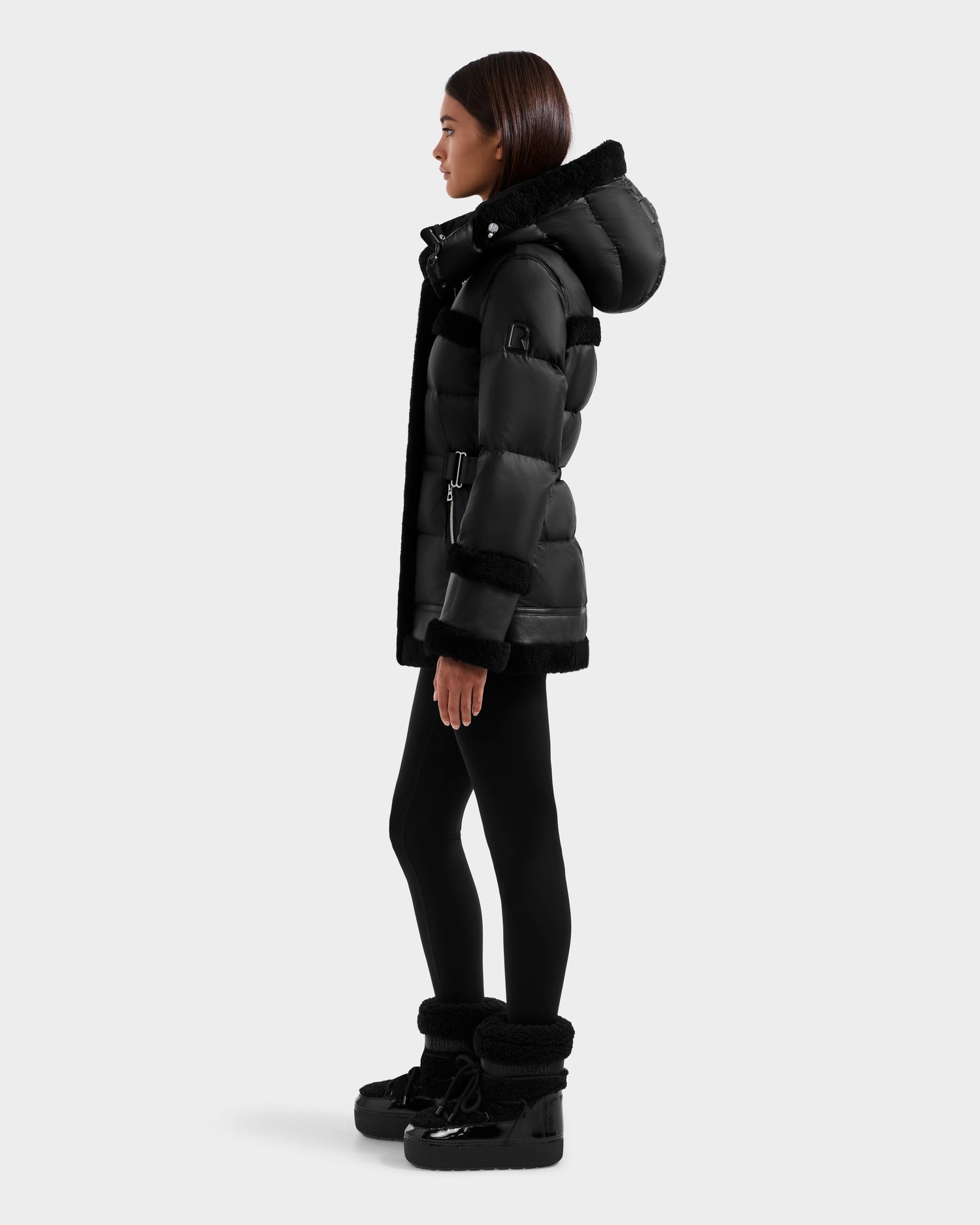 A woman stands in profile wearing the Rudsak TESSA - 8125615 Black down puffer jacket with a hood, black leggings, and black ankle boots with shearling trim against a plain light gray background.