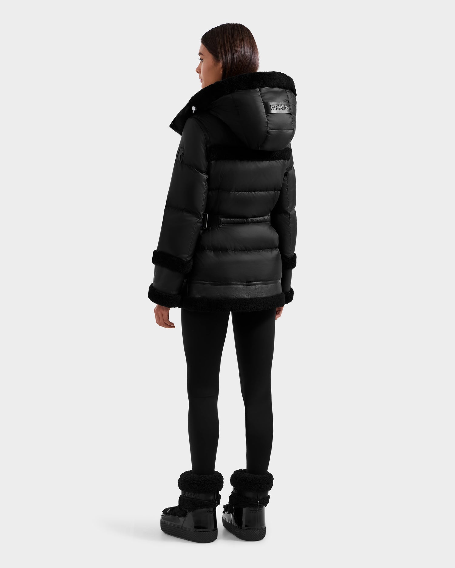 A woman faces away, wearing the Rudsak TESSA - 8125615 Black women's down puffer jacket with a fur-lined hood, black leggings, and black fur-trimmed boots against a plain white background.