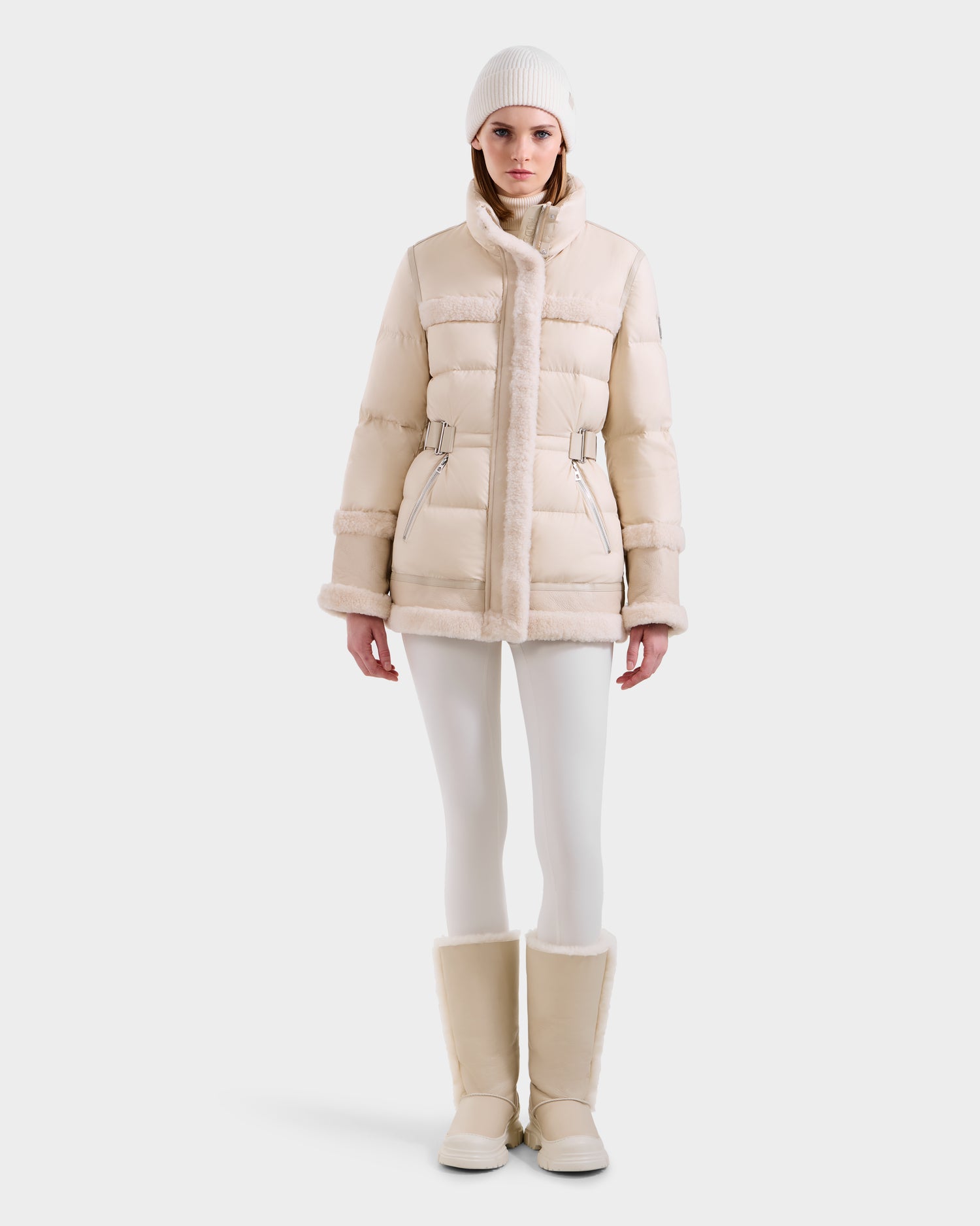 A person wearing the Rudsak TESSA - 8125615 Vanilla women’s down puffer with shearling trim, fitted pants, fur-lined boots, and a knit beanie stands facing forward against a plain white background, showcasing a chic winter look.