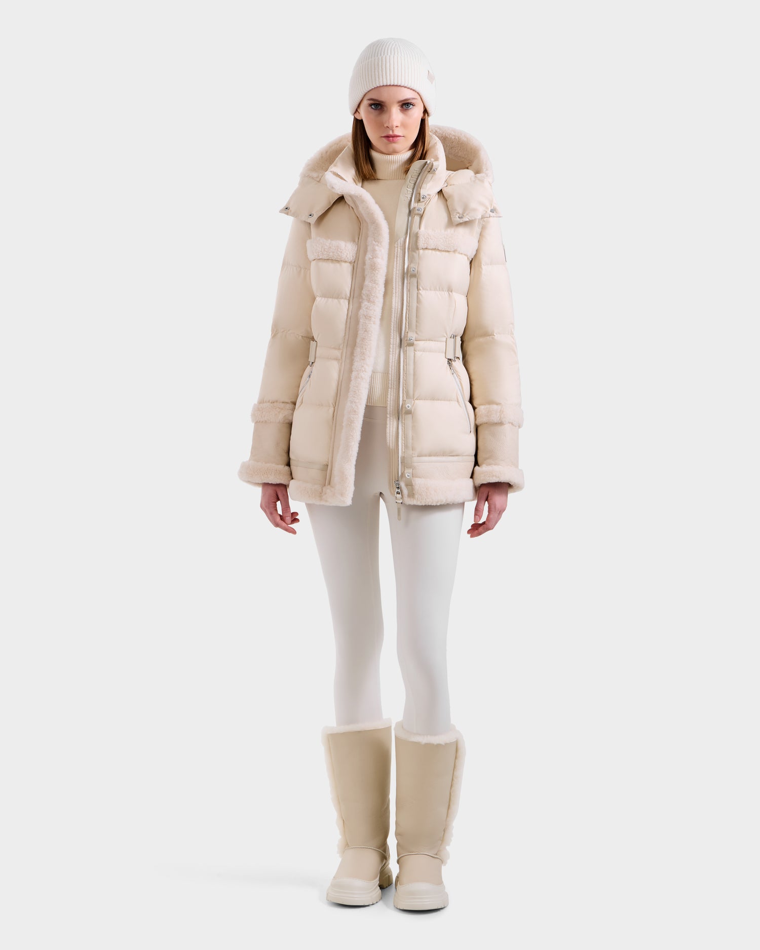 A woman in the Rudsak TESSA - 8125615 Vanilla down puffer with shearling trim, white beanie, turtleneck, leggings, and fur-lined boots stands facing forward against a white background—a true winter essential look.