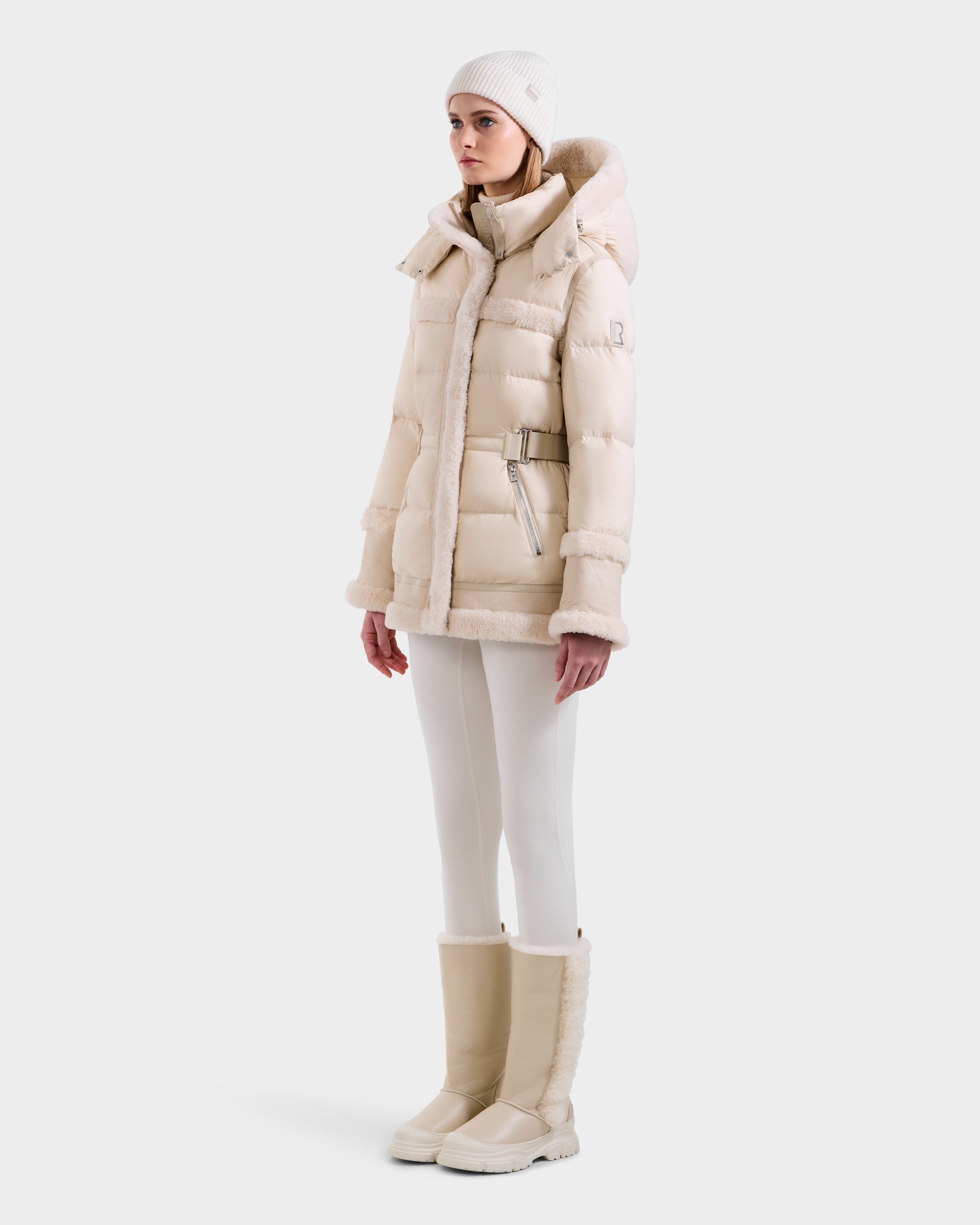 A woman models the Rudsak TESSA - 8125615 Vanilla down puffer with belt, paired with fitted pants, a knit beanie, and shearling-trimmed tall boots—capturing an essential winter look against a plain white background.