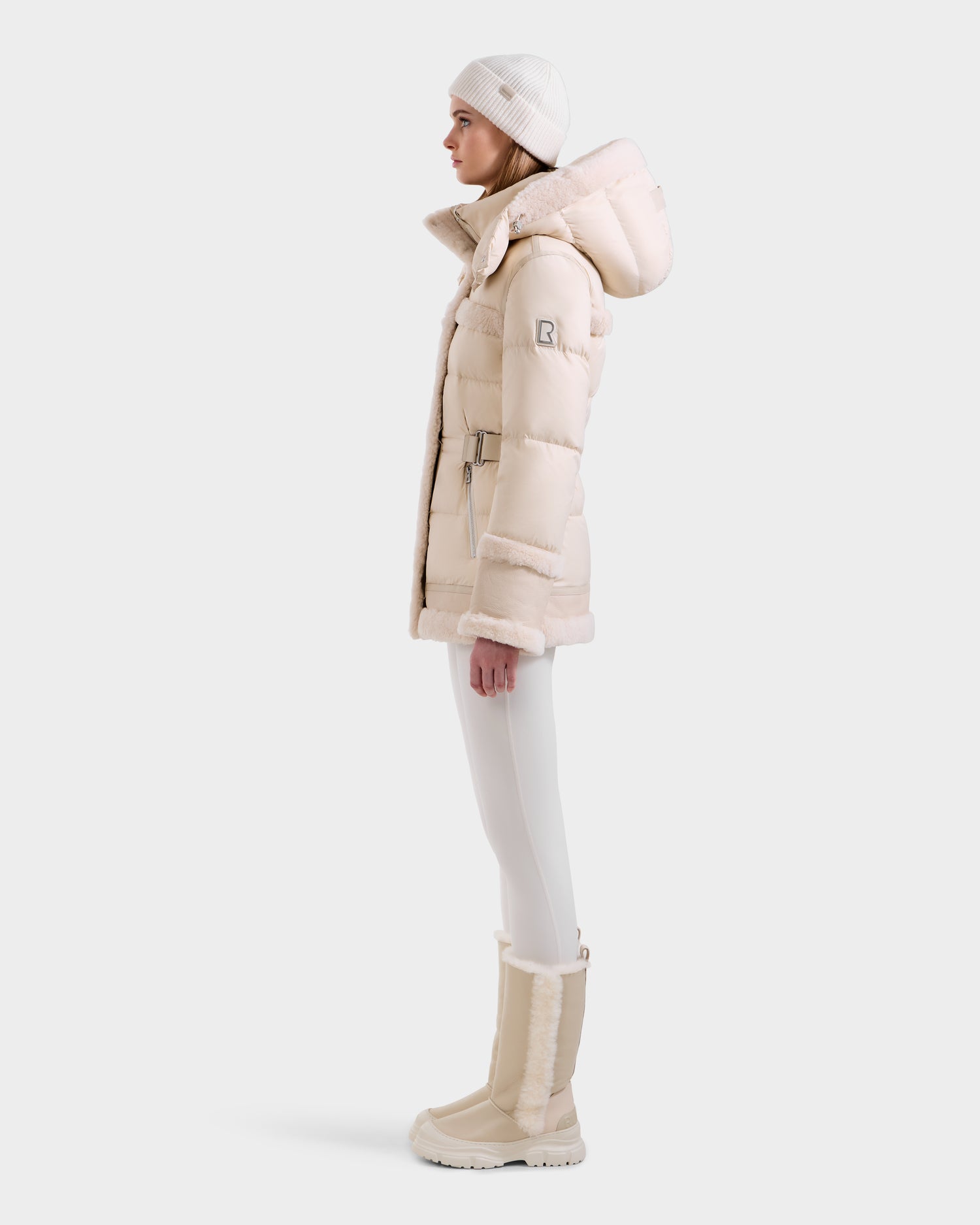 A woman in profile wears the Rudsak TESSA - 8125615 Vanilla down puffer jacket with a hood, a matching knit hat, white leggings, and beige winter boots—a must-have for winter—against a plain white background.