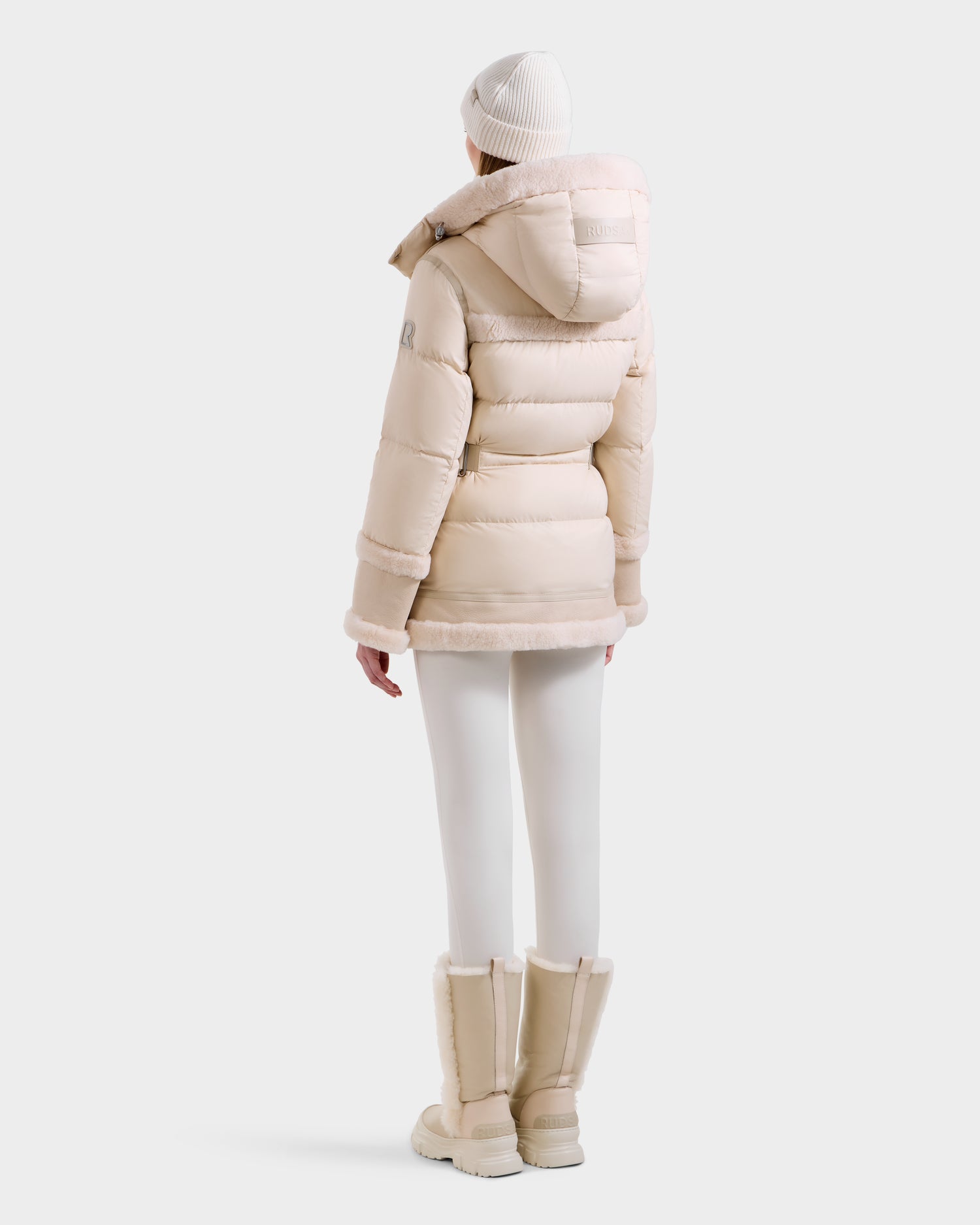 A woman wearing the Rudsak TESSA - 8125615 Vanilla down puffer jacket with hood, white leggings, tall beige winter boots, and a white knit beanie stands facing away against a plain light background.
